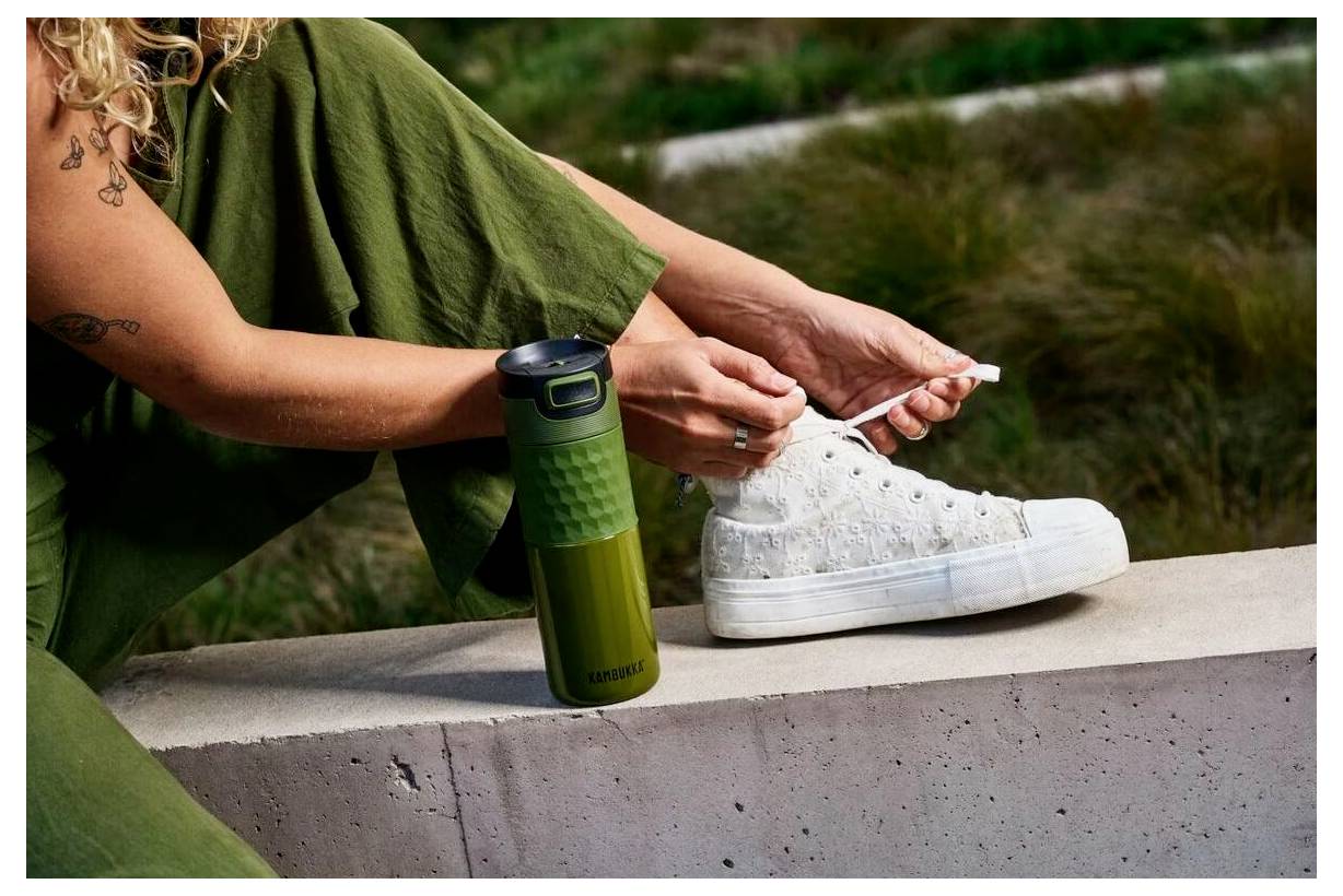 Eine Person in grüner Hose bindet weiße Sneaker auf einer Betonkante neben einer grünen Thermoskanne an, mit Gras im Hintergrund.