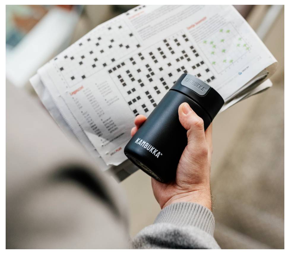 Eine Person hält einen Reisebecher, während sie eine Zeitung mit Sudoku-Rätseln und einem Punktdiagramm liest und dabei Freizeit und Entspannung betont.