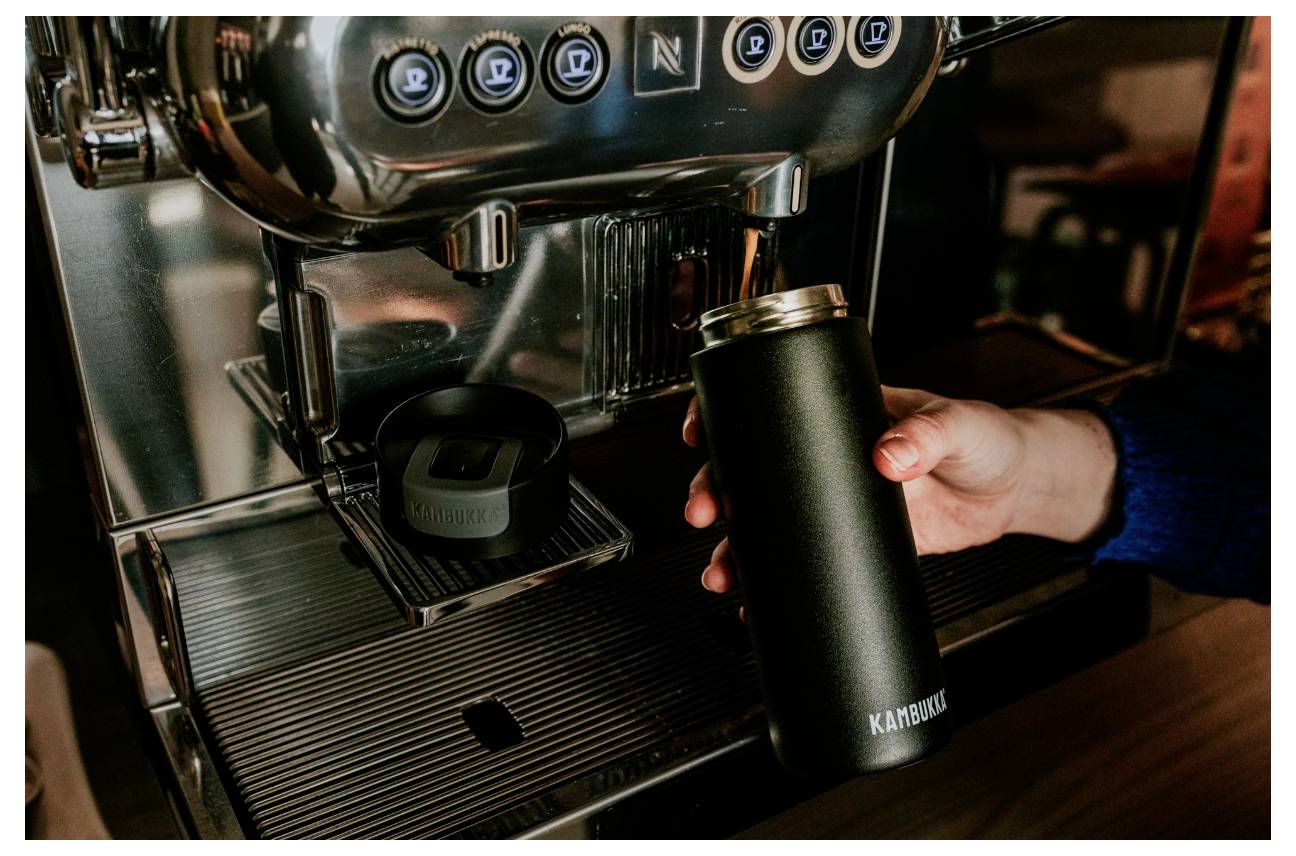 Eine Person hält eine schwarze Thermoskanne unter einer Kaffeemaschine und bereitet sich darauf vor, diese zu füllen. Knöpfe an der Maschine zeigen verschiedene Getränkeoptionen an.