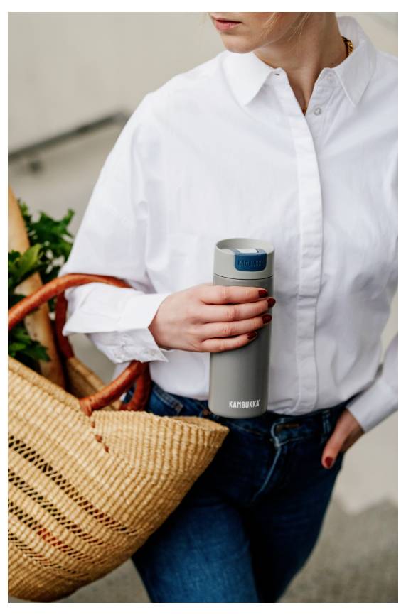 Eine Person in einem weißen Hemd hält eine graue wiederverwendbare Wasserflasche und einen geflochtenen Korb mit Gemüse.