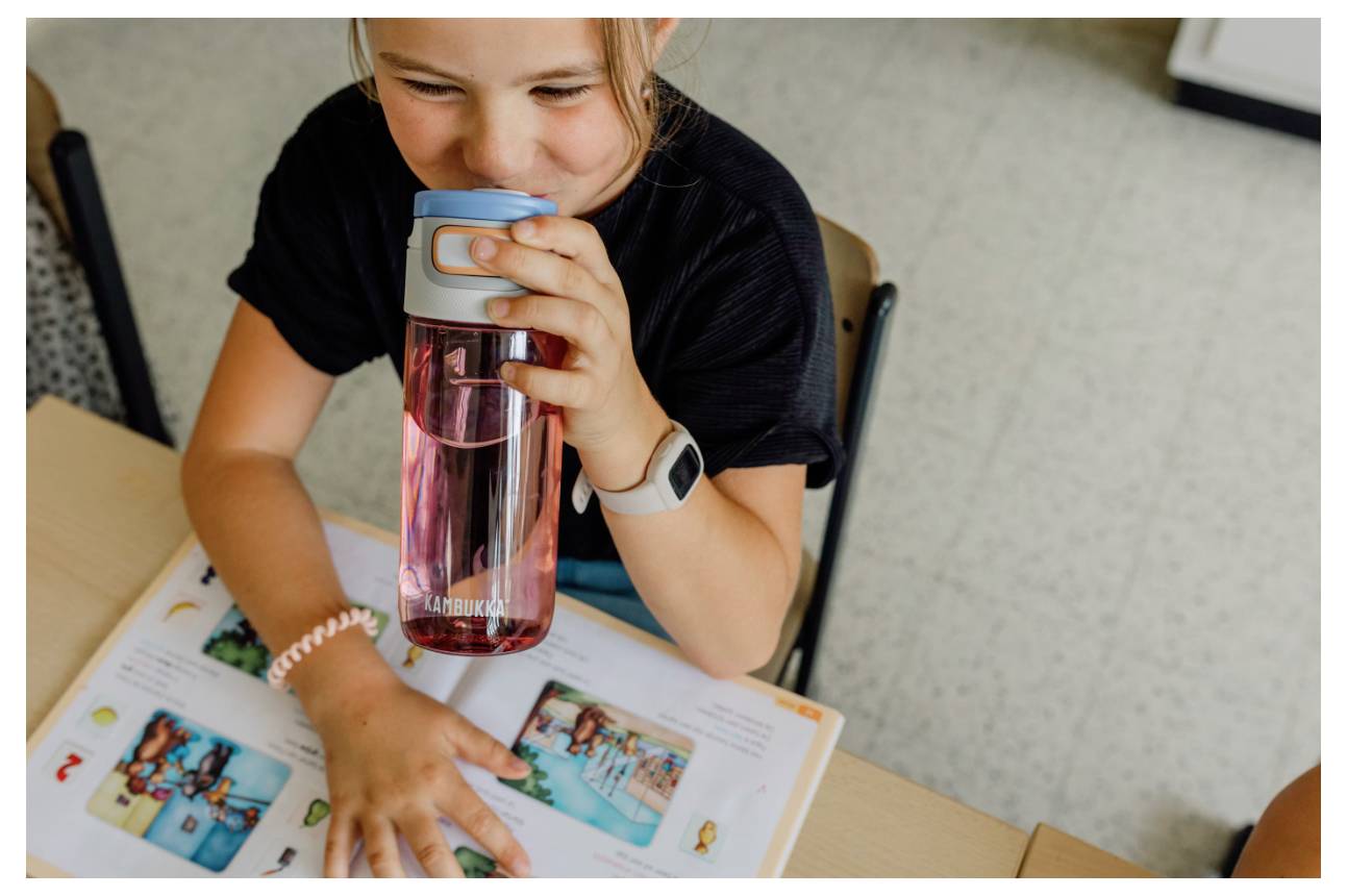 Ein Kind lächelt, während es aus einer rosa Wasserflasche trinkt, sitzt an einem Schreibtisch und schaut auf ein buntes aufgeschlagenes Buch.