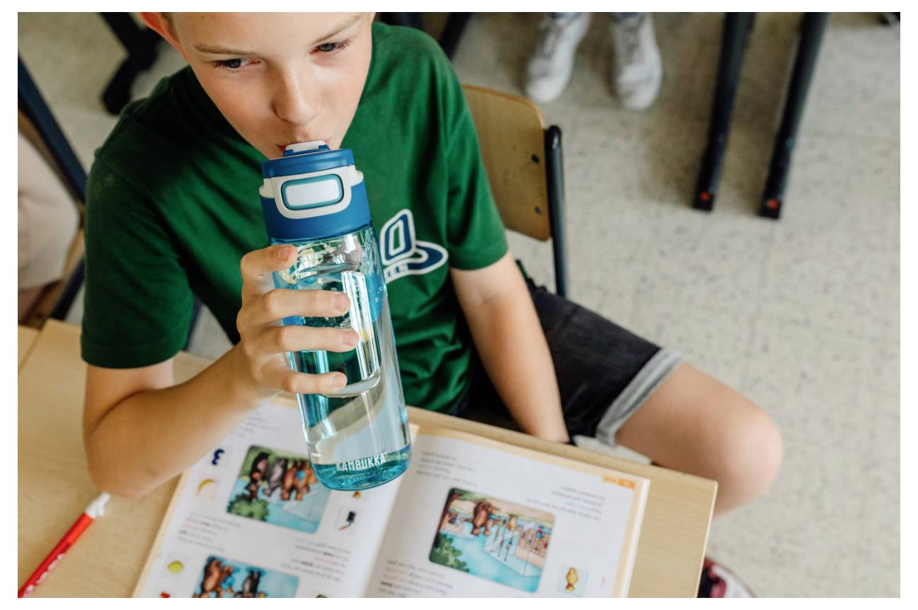 Ein Kind in einem grünen Hemd sitzt an einem Schreibtisch und trinkt aus einer blauen Wasserflasche, mit einem aufgeschlagenen illustrierten Buch auf dem Schreibtisch.