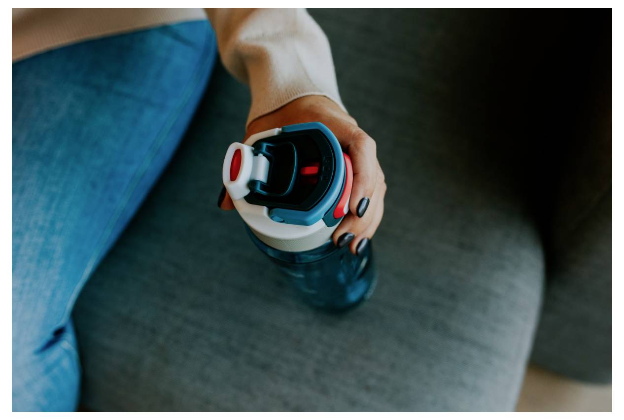 Eine Person hält eine Wasserflasche, während sie auf einem grauen Sofa sitzt, und zeigt ihre lackierten Nägel und ihre legere Kleidung.