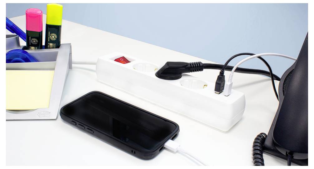 Ein Smartphone, das von einer Steckdosenleiste auf einem Schreibtisch aufgeladen wird, neben Büromaterialien wie Textmarkern, Klebeband und Haftnotizen.