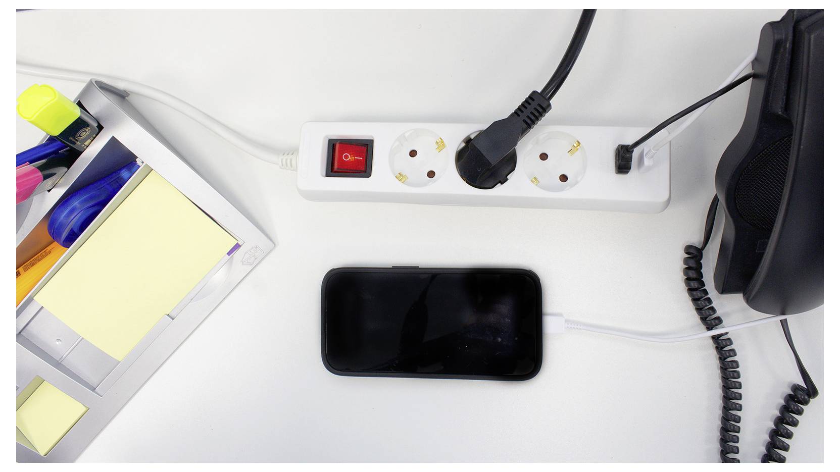 Ein Smartphone lädt auf einem Schreibtisch auf und ist mit einer Steckerleiste mit einem roten Schalter verbunden. In der Nähe befinden sich Büromaterialien und ein Telefon.