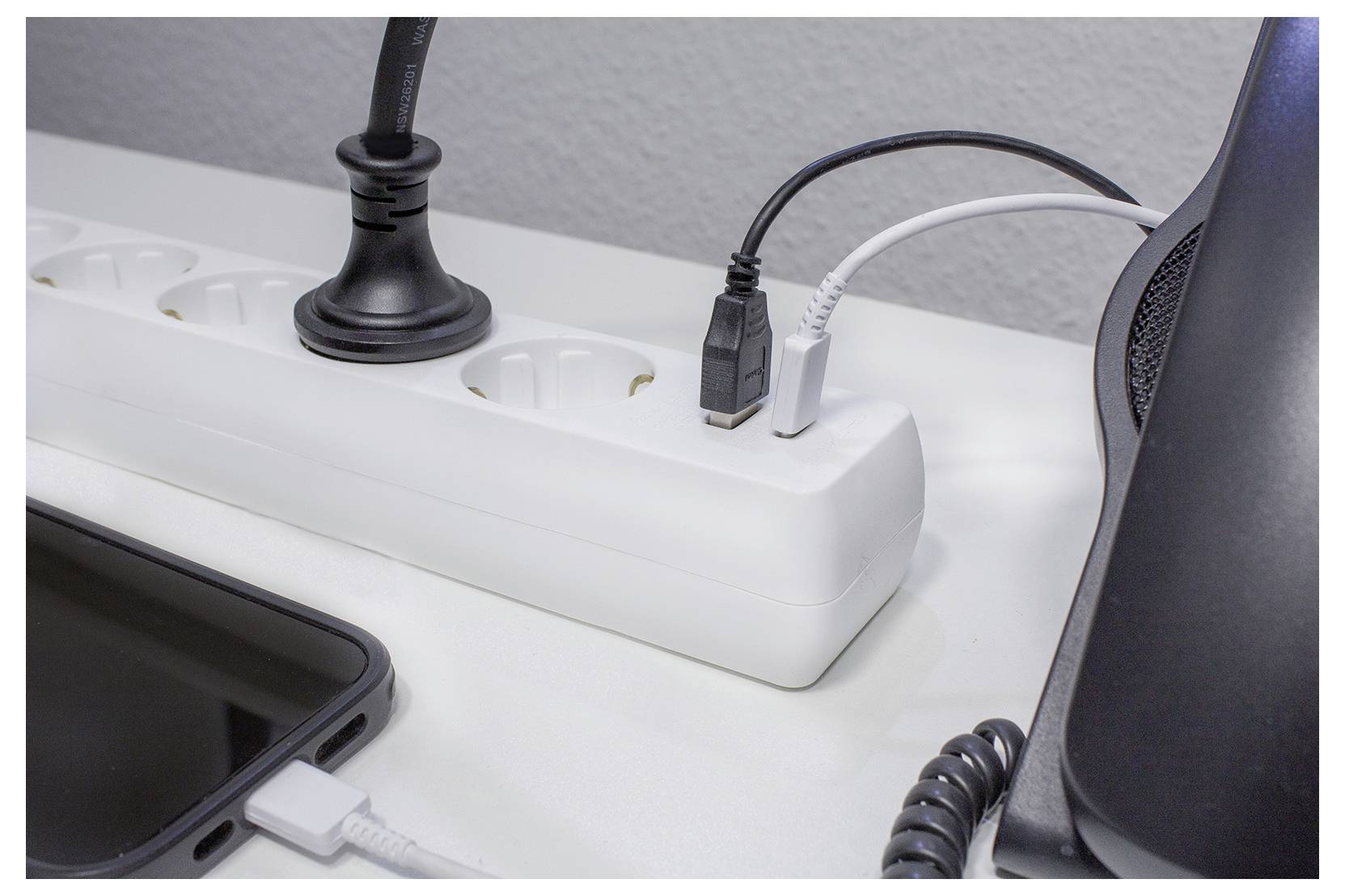 Steckerleiste auf einem Tisch mit mehreren Steckdosen, einschließlich eines angeschlossenen schwarzen Stromkabels und eines angebrachten weißen USB-Kabels, in der Nähe eines Telefons und eines Lautsprechers.