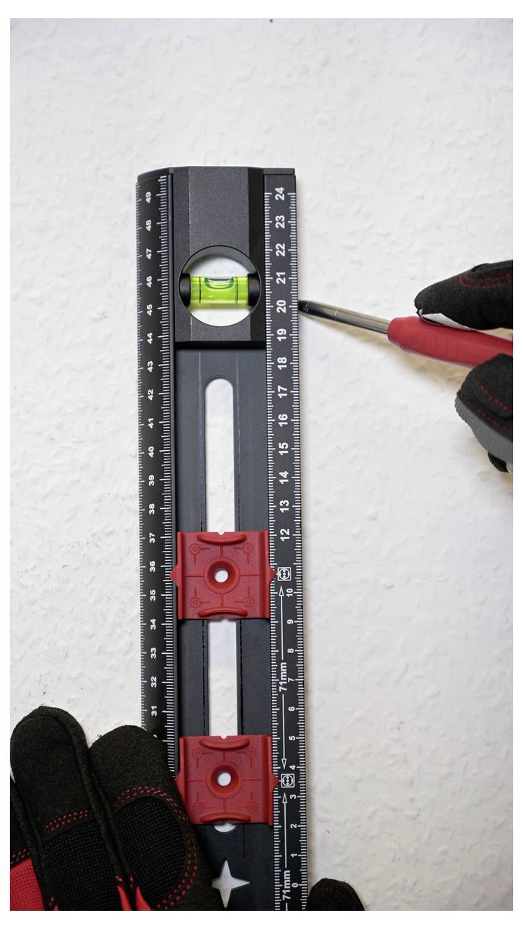 A gloved hand holds a black level tool with a bubble vial against a wall, while another hand marks a spot with a red-handled screwdriver.