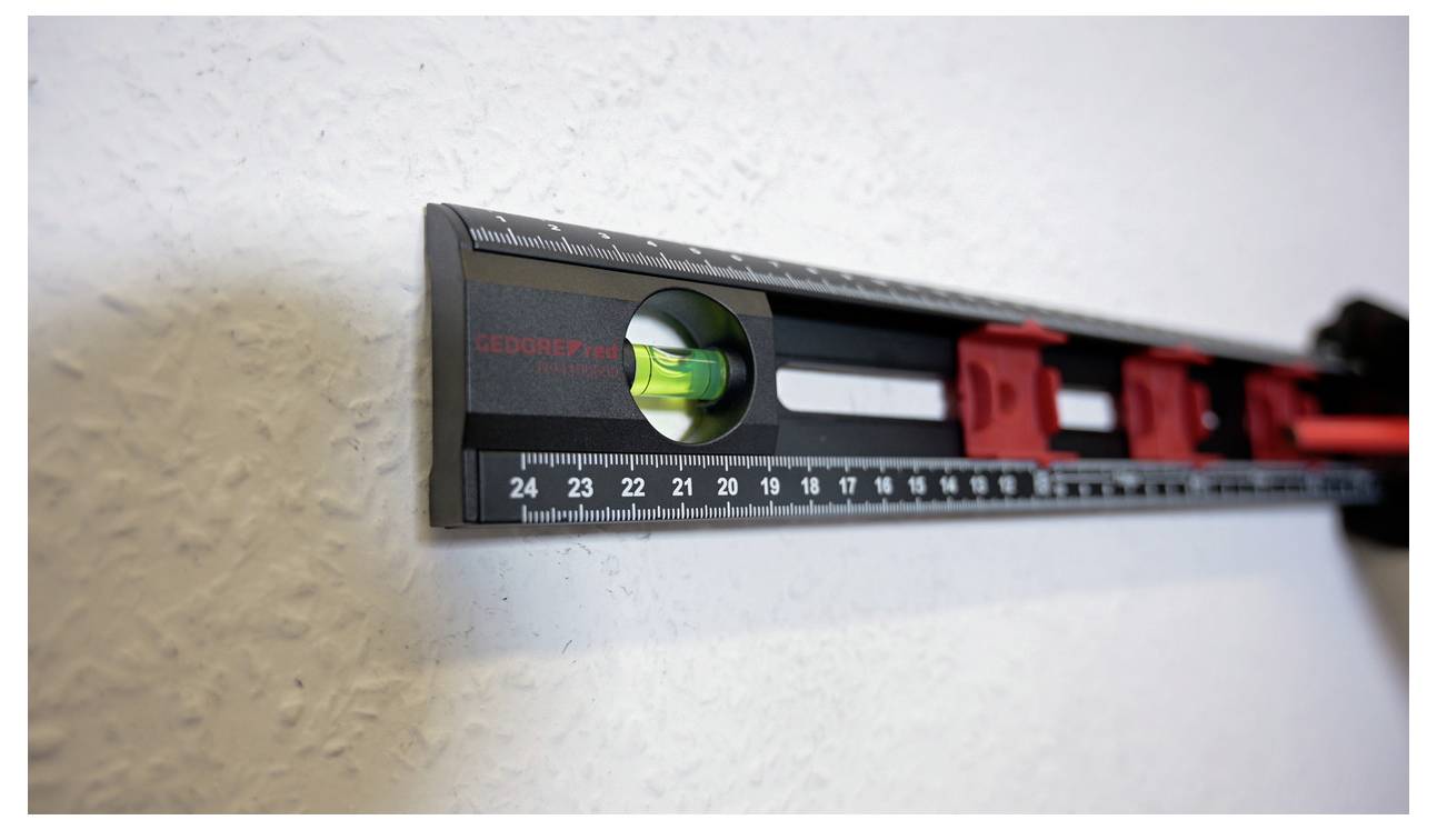 A bubble level on a black and red ruler is positioned horizontally on a white wall, showing that the surface is level.