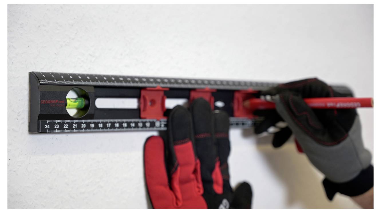 A person wearing gloves uses a black and red level tool and pencil to mark a wall for alignment.