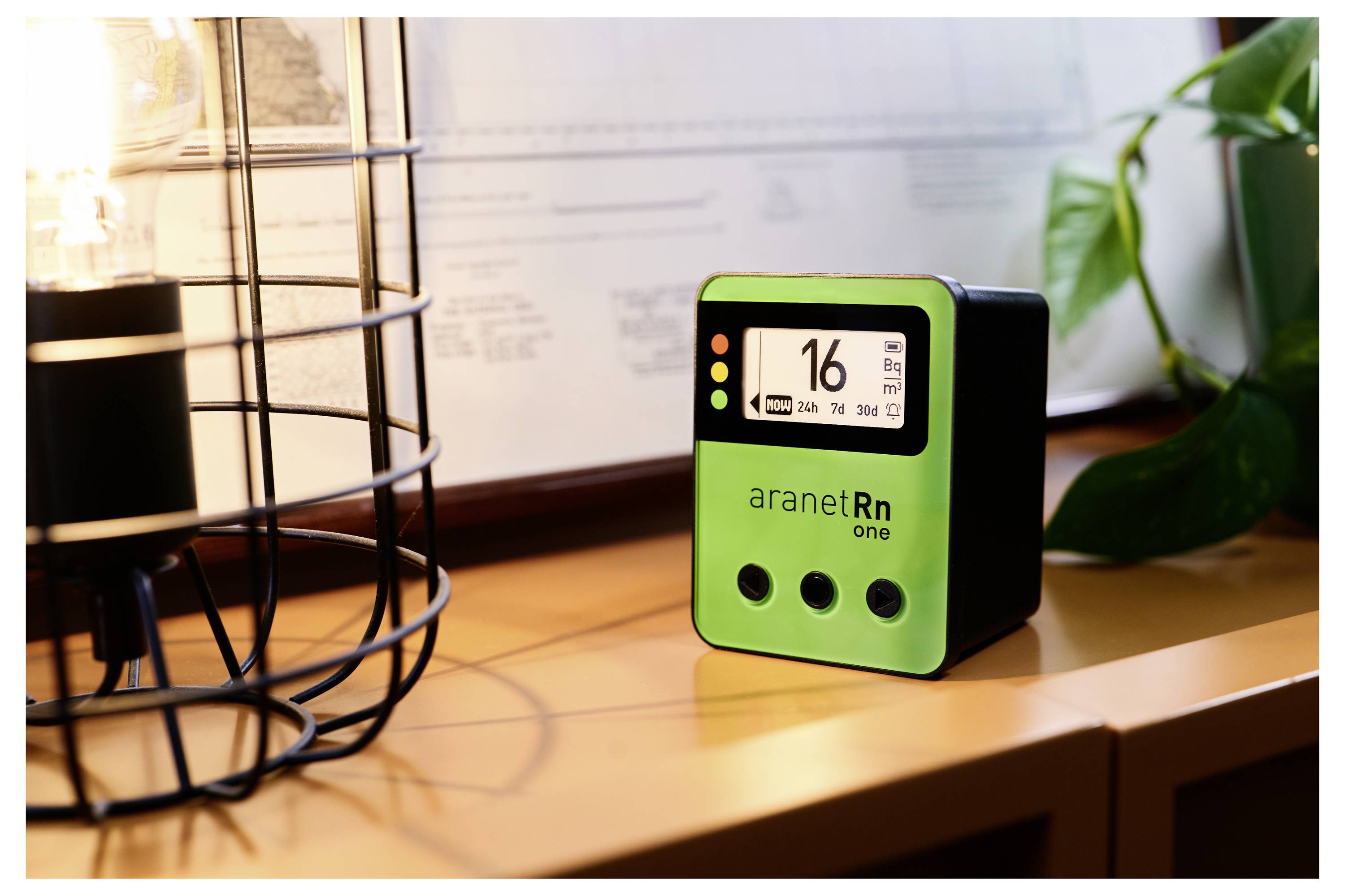 Ein grünes Radon-Messgerät auf einem Tisch zeigt '16 Bq/m³' an. Im Hintergrund sind ein Licht und eine Pflanze zu sehen.