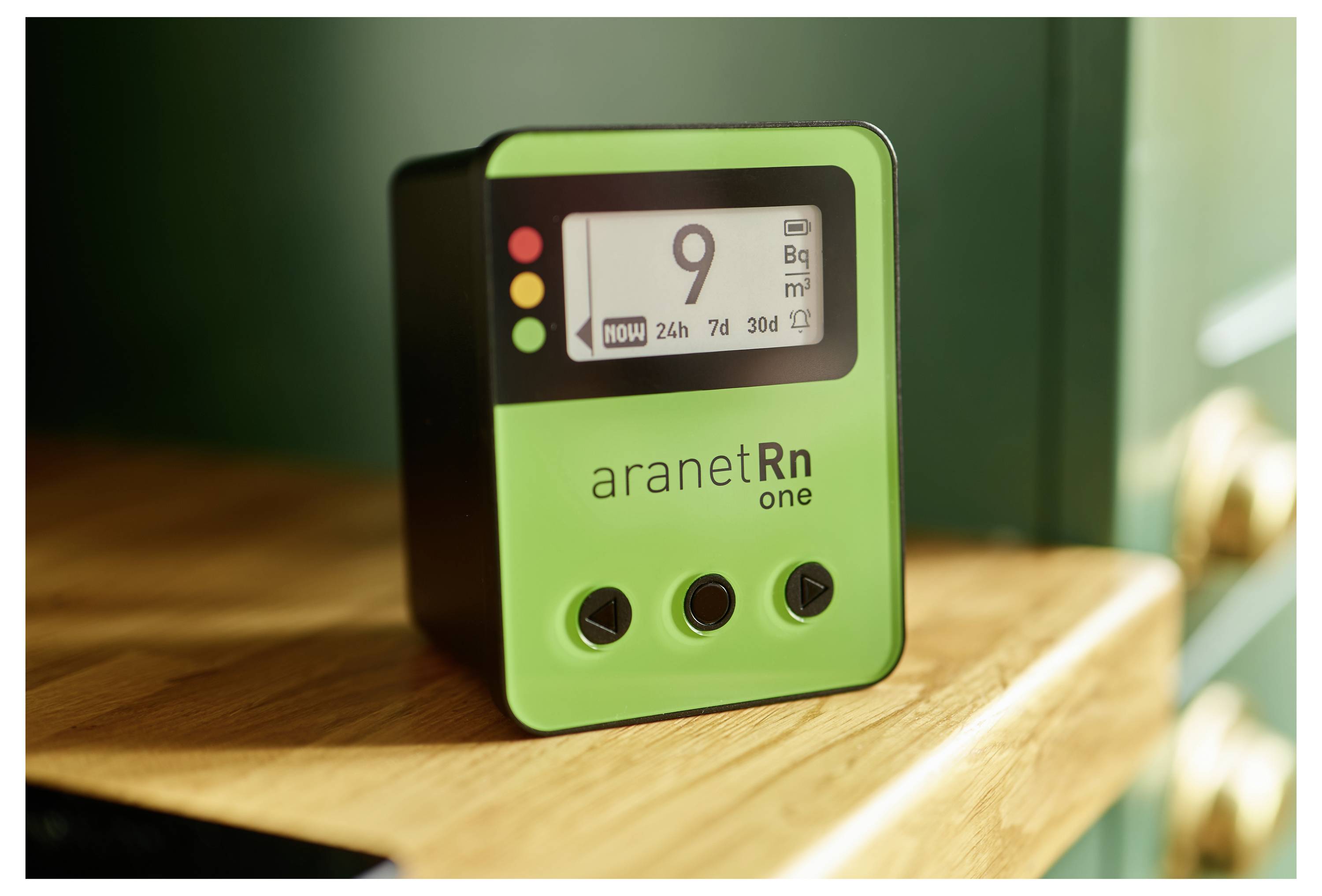 Ein grünes Gerät zeigt die Radonkonzentration an einem Bildschirm an. Es steht auf einem Holzregal und hat drei Bedienknöpfe.