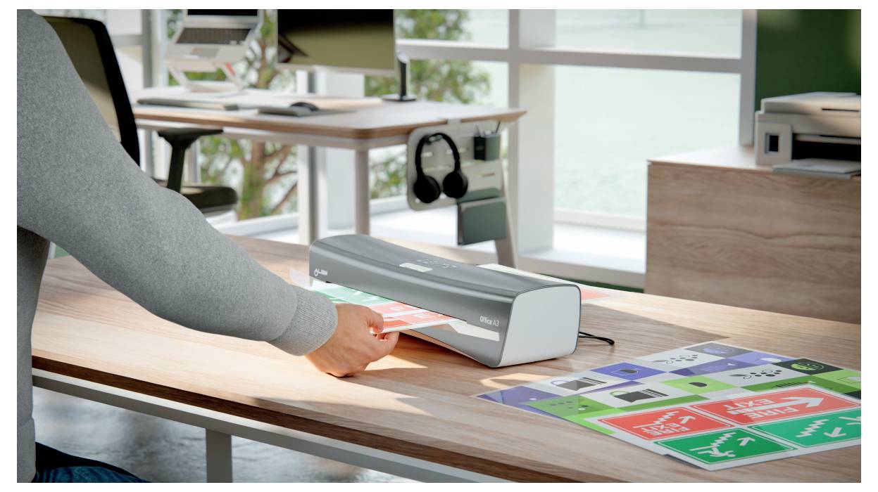 Eine Person laminiert bunte Sicherheitsschilder mit einem Laminiergerät auf einem Holzschreibtisch in einem gut beleuchteten Büro mit großen Fenstern im Hintergrund.