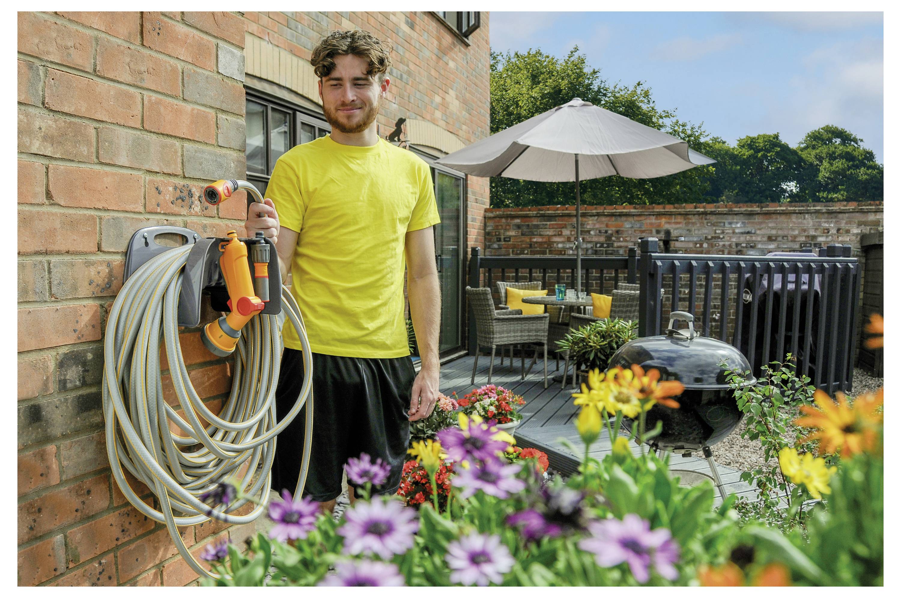 Mann in gelbem Hemd, der eine Schlauchrolle hält, steht in einem Hinterhof mit Blumen, Terrassenmöbeln und einem Grillgerat in der Nähe.