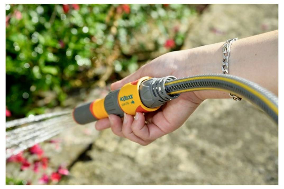 Eine Person, die einen Gartenschlauch hält und Blumen entlang eines gepflasterten Weges bewässert, mit Fokus auf die Schlauchöse und das Wasserspritzmuster.