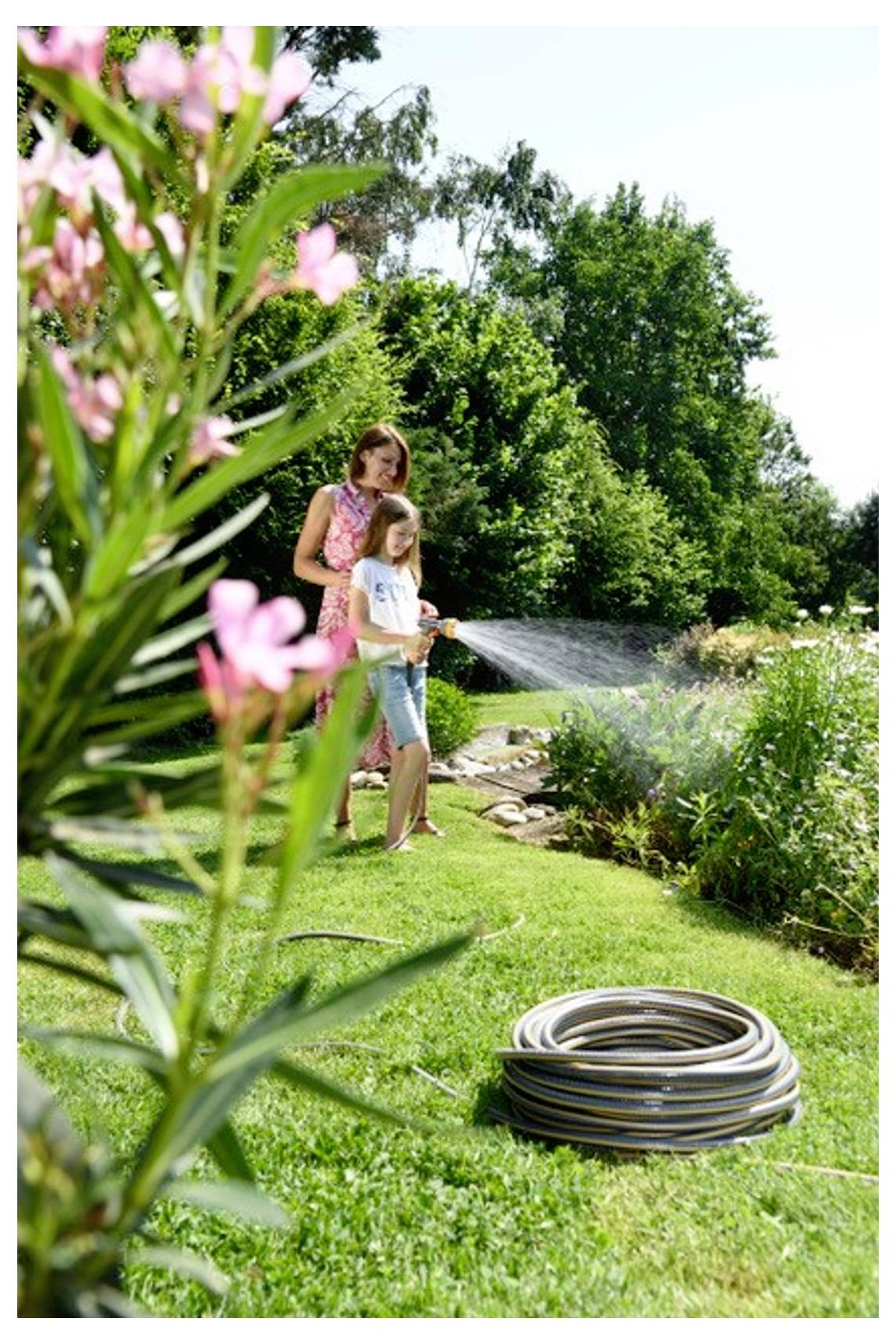 Mutter und Kind gießen den Garten an einem sonnigen Tag mit einem Schlauch, umgeben von Grünflächen und blühenden Blumen.
