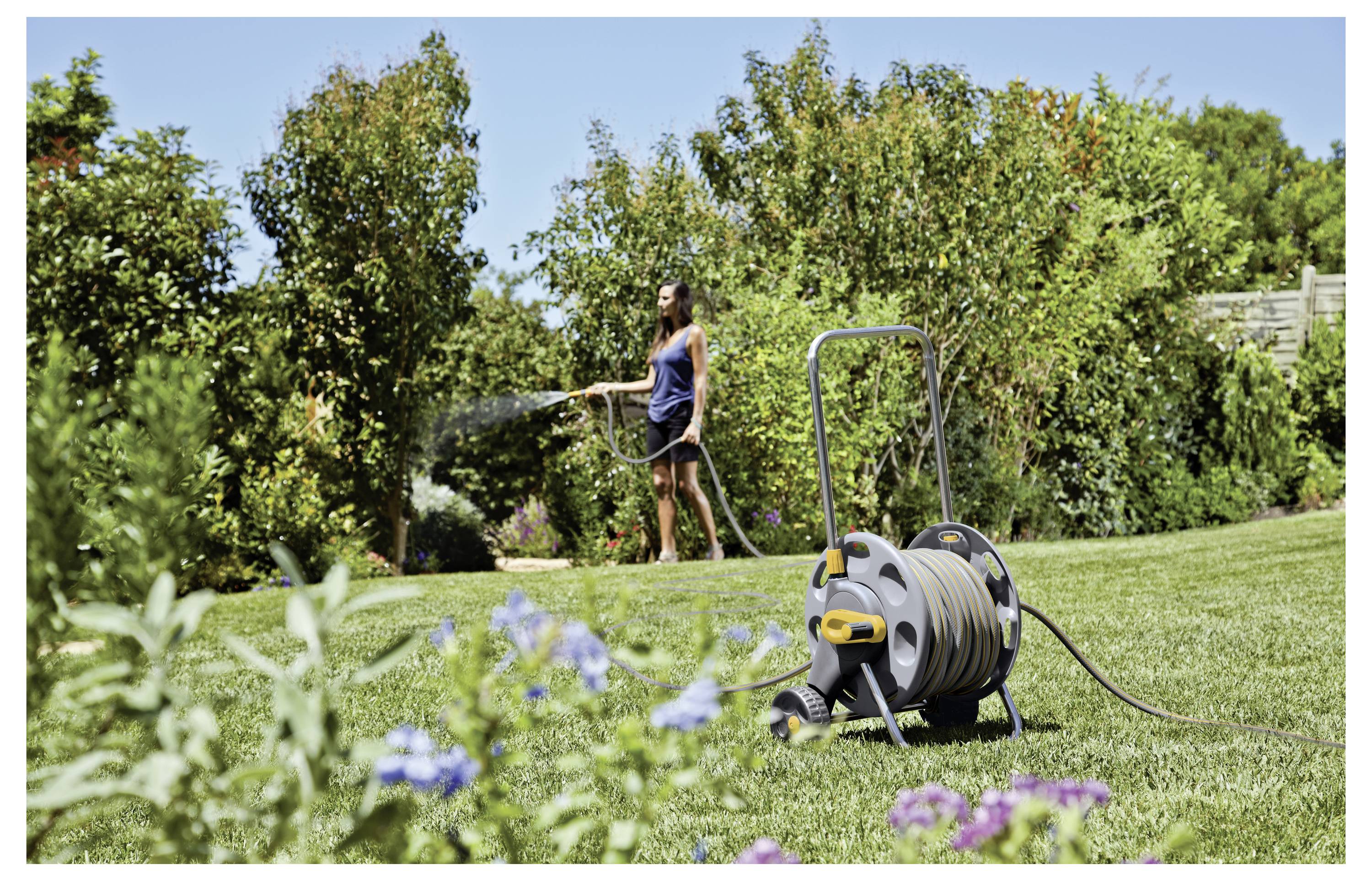 Eine Frau bewässert einen Garten mit einem Schlauch, der an einer Spule auf einer Rasenfläche befestigt ist, umgeben von grünen Bäumen und blühenden Pflanzen.