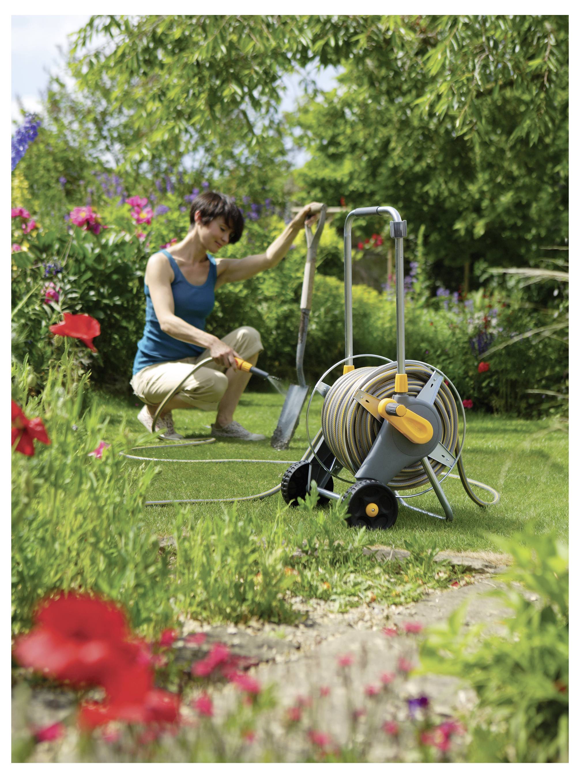 Eine Frau, die in einem farbenfrohen Blumengarten mit einer Schlauchrolle gärtnert, umgeben von verschiedenen blühenden Pflanzen und Grün.