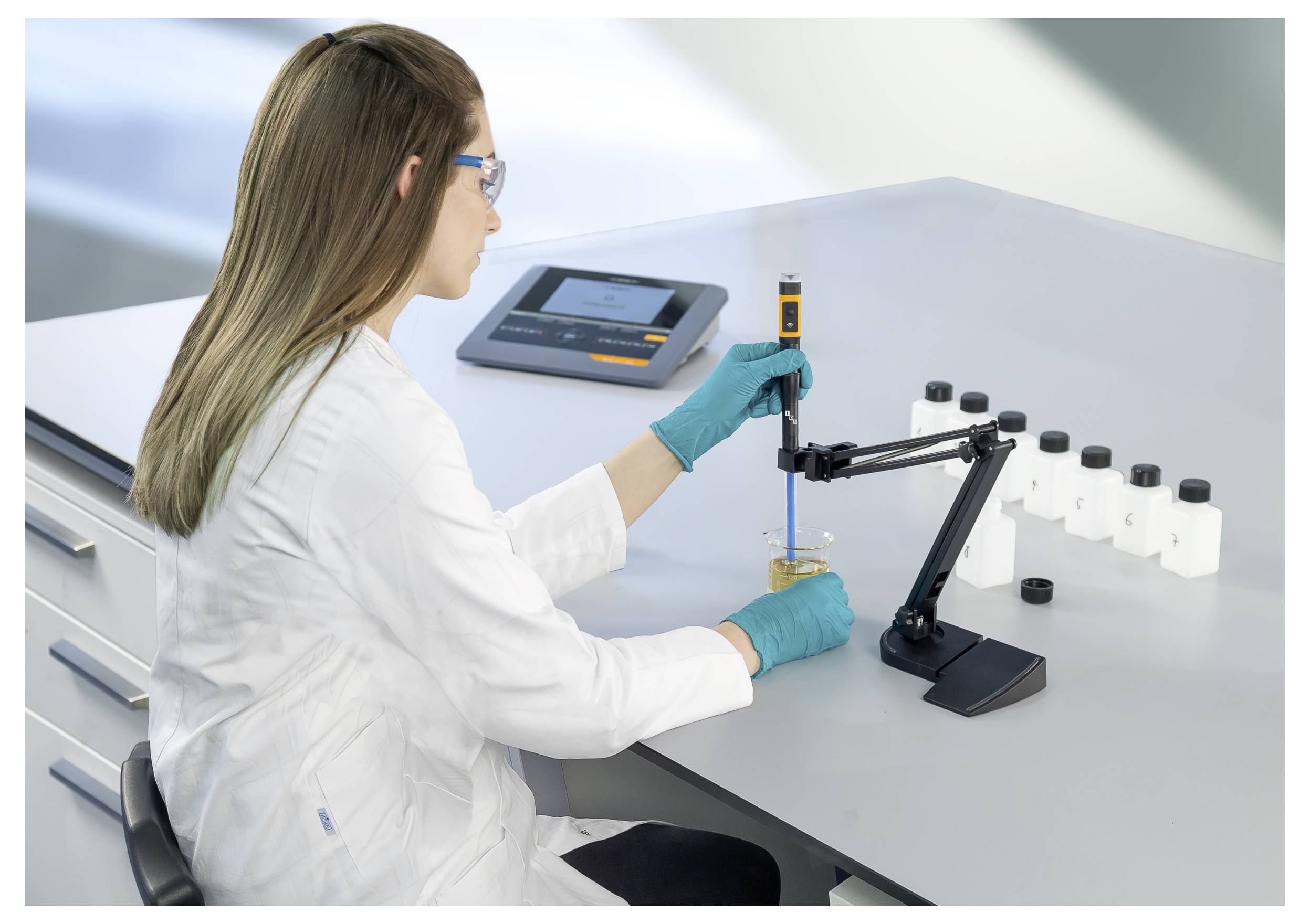 Ein Wissenschaftler in einem Laborkittel verwendet eine digitale Pipette an einer Probe. Auf dem Tisch befinden sich Flaschen und ein Gerät, im Hintergrund befindet sich Laborausrüstung.