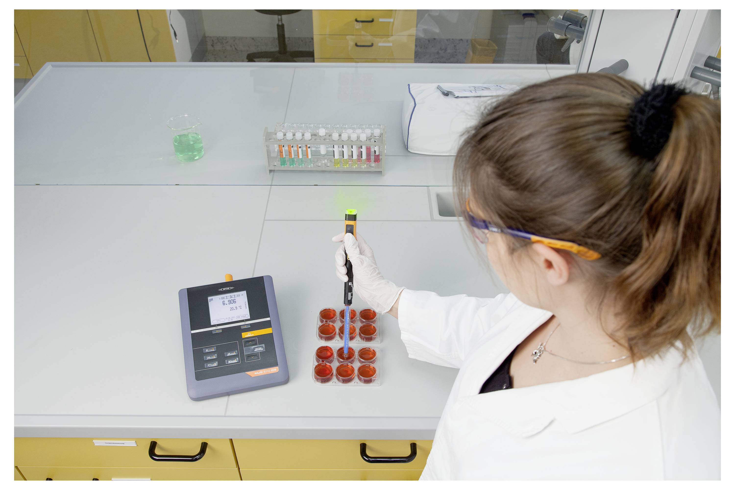 Ein Wissenschaftler im Laborkittel verwendet eine Pipette auf Petrischalen, die auf einem Labortisch platziert sind. Eine digitale Waage und farbige Proben sind in der Nähe sichtbar.