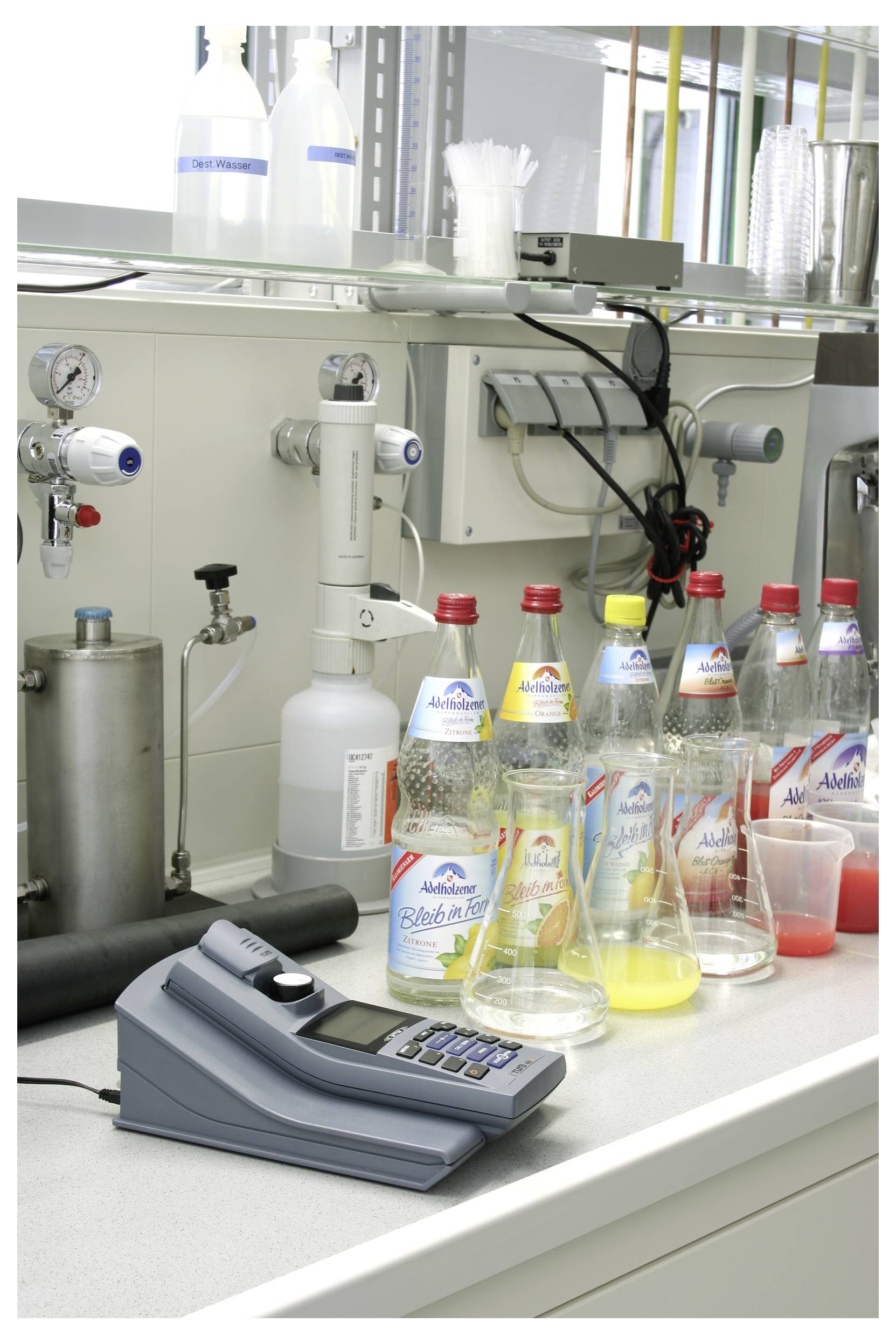 Labortisch mit verschiedenen Adelholzener Mineralwasserflaschen, einem Messinstrument und Laborausrüstung, was auf Tests oder Analysen hindeutet.
