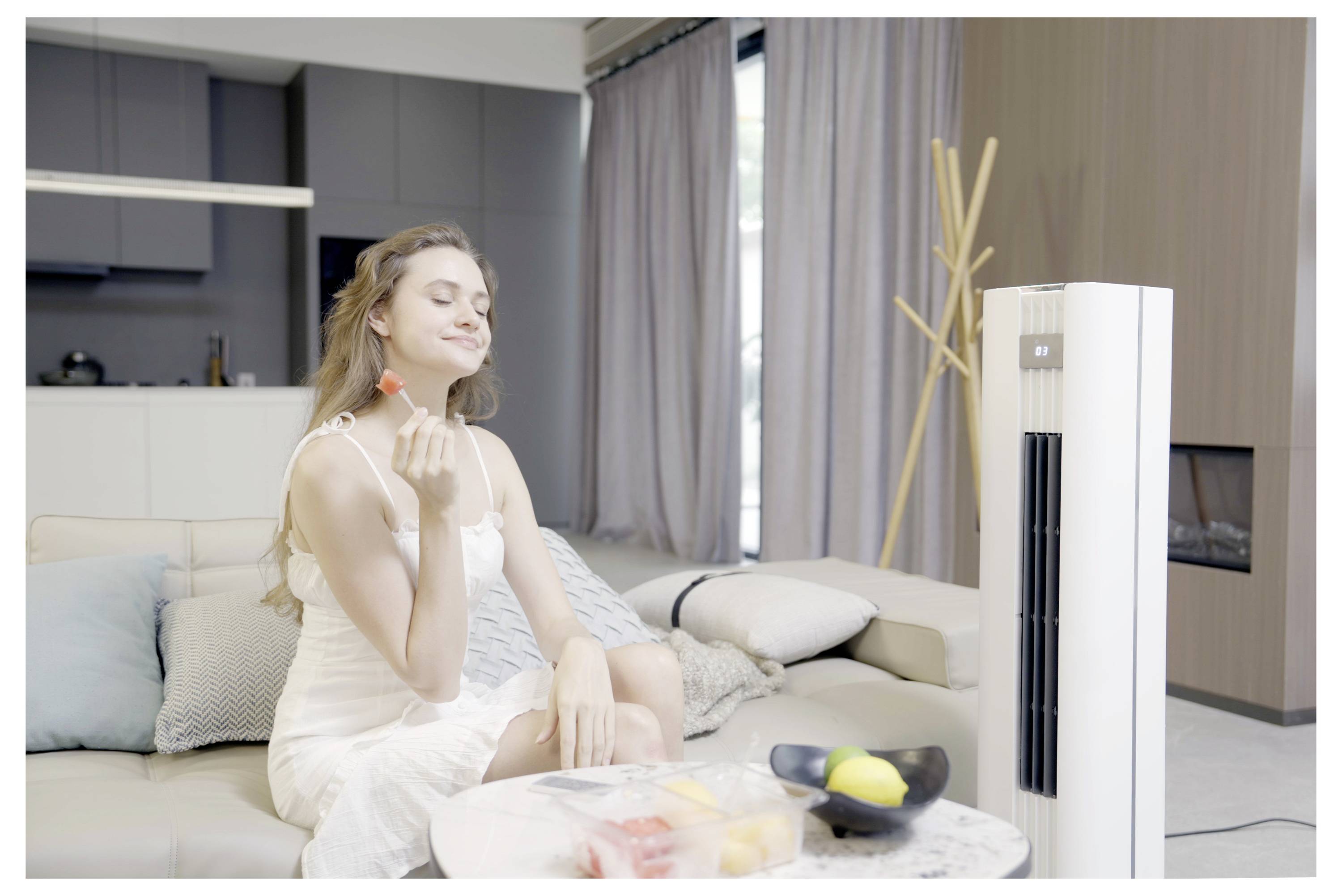 Eine Frau in einem weißen Kleid genießt ein Stück Wassermelone auf einem Sofa und sitzt in einem modernen Wohnzimmer mit einem Luftreiniger in der Nähe.