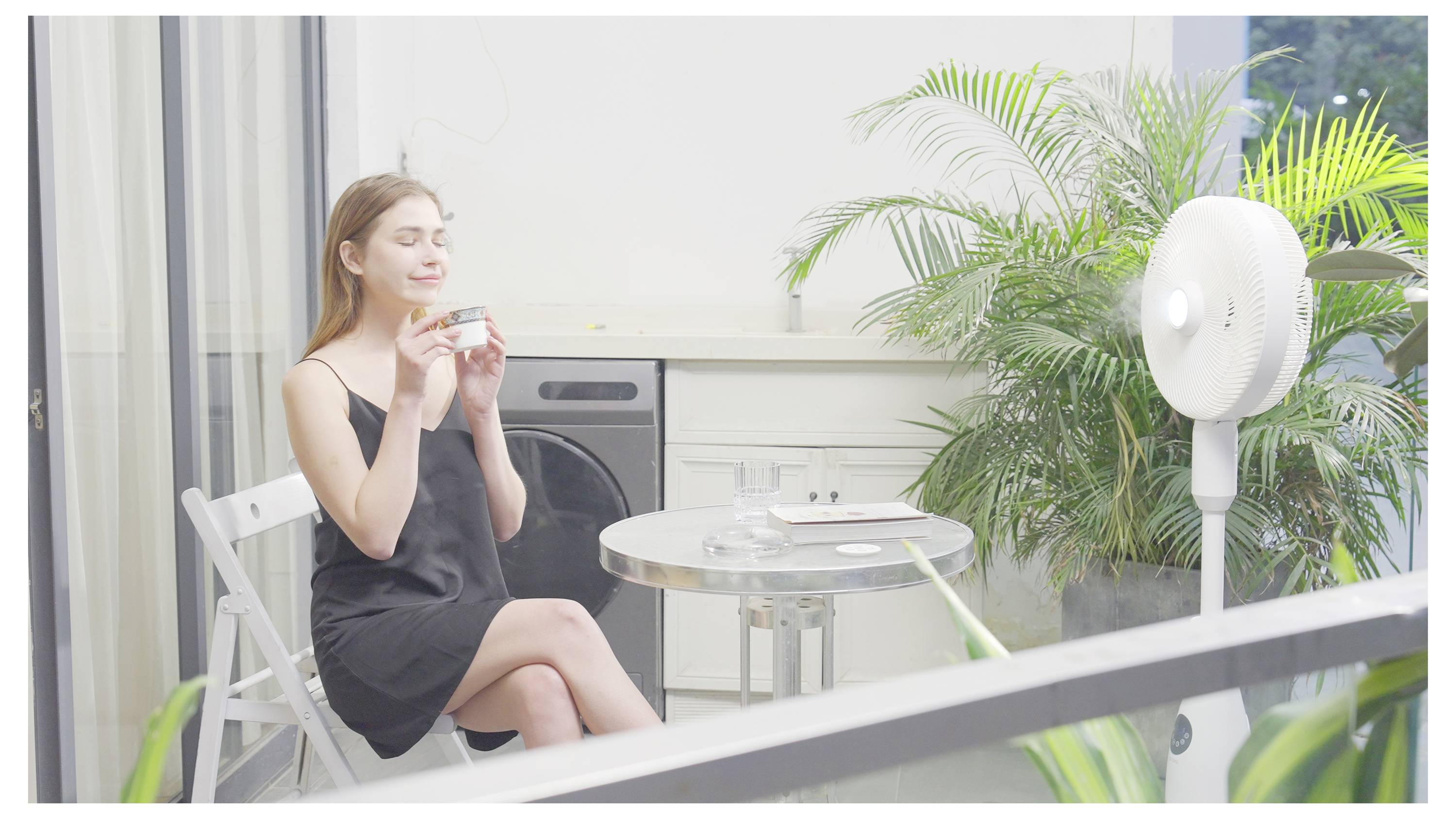 Eine Frau in einem schwarzen Kleid sitzt an einem kleinen Tisch in einem hellen Raum und hält ein Glas in der Hand, während sie lächelt. Ein Ventilator und grüne Pflanzen sind in der Nähe.