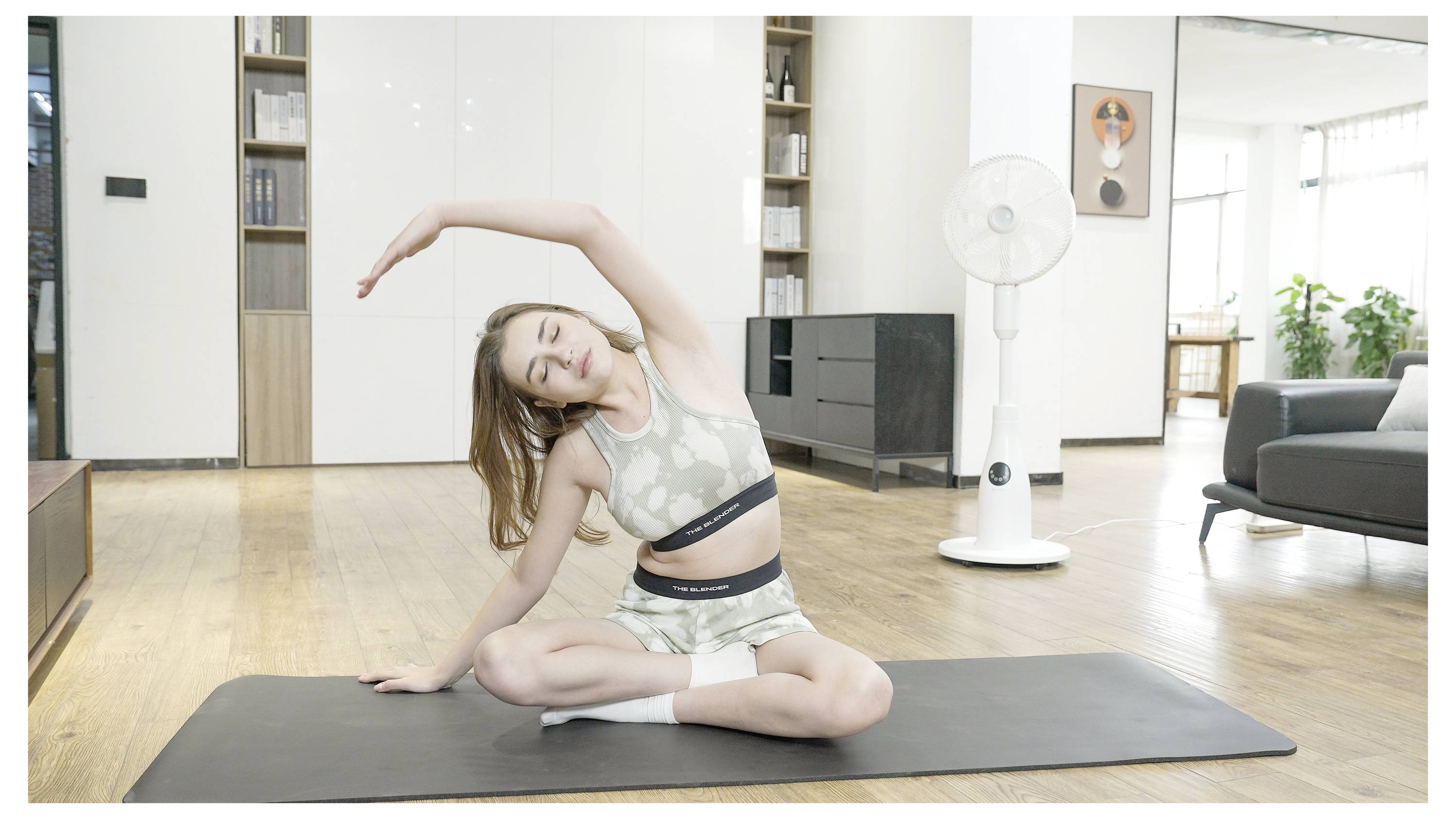 Eine Frau in Sportkleidung führt eine Seitstreckung auf einer Yogamatte in einem modernen Wohnzimmer mit einem Ventilator und einem Bücherregal im Hintergrund durch.