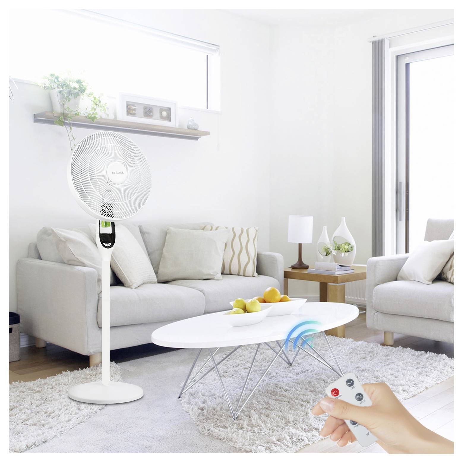 Ein modernes Wohnzimmer mit einem weißen Standventilator, ferngesteuert, in der Nähe von Sofas und einem Couchtisch mit Obst. Eine Hand hält die Fernbedienung.