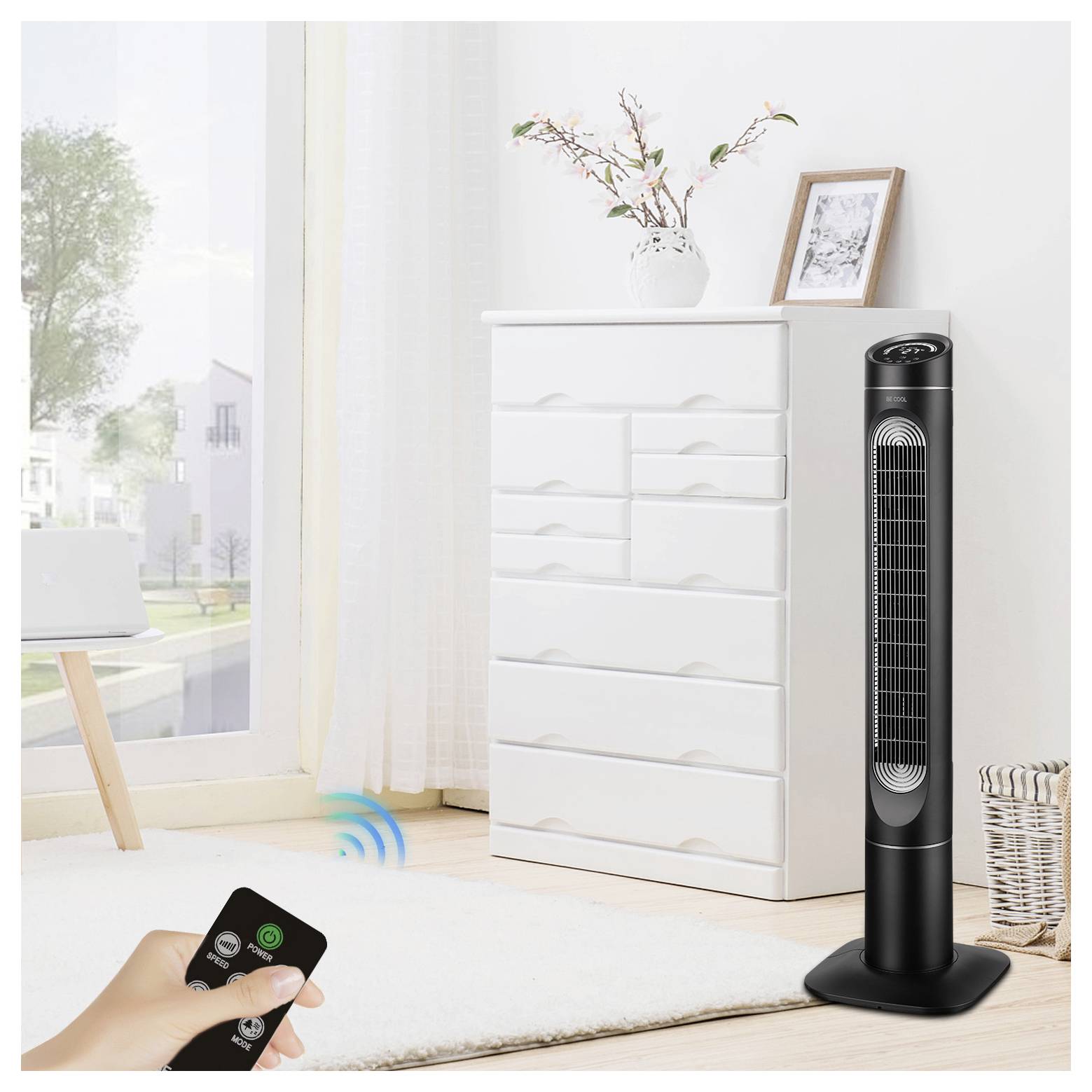 Eine Person verwendet eine Fernbedienung, um einen großen, schwarzen Turmventilator in einem hellen Raum mit einer weißen Kommode, einer Pflanze und einem gerahmten Foto obenauf zu steuern.