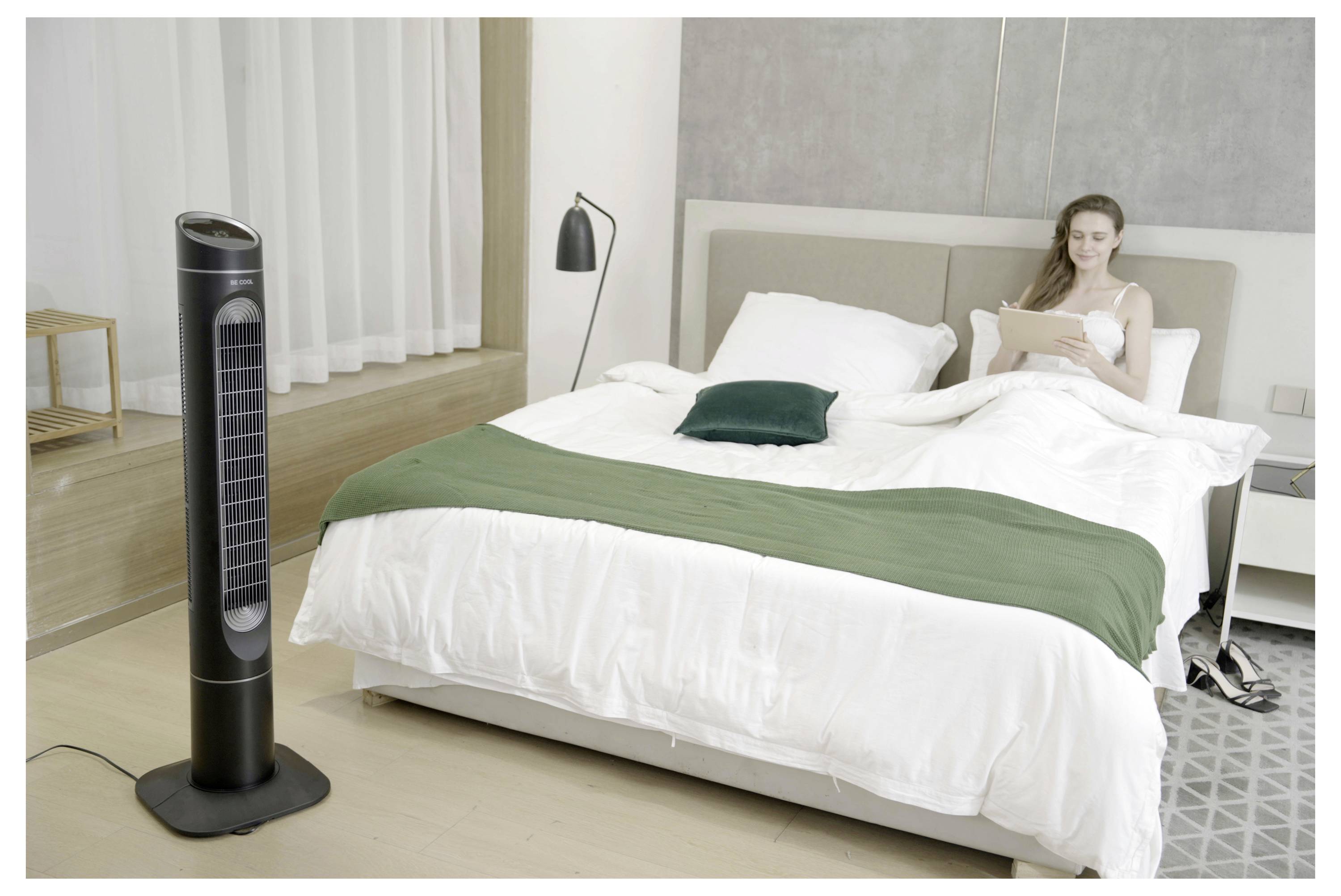 Eine Frau sitzt auf einem Bett in einem hellen, modernen Schlafzimmer und liest auf einem Tablet. Ein großer schwarzer Ventilator steht in der Nähe und deutet auf einen Fokus auf Komfort und Entspannung hin.