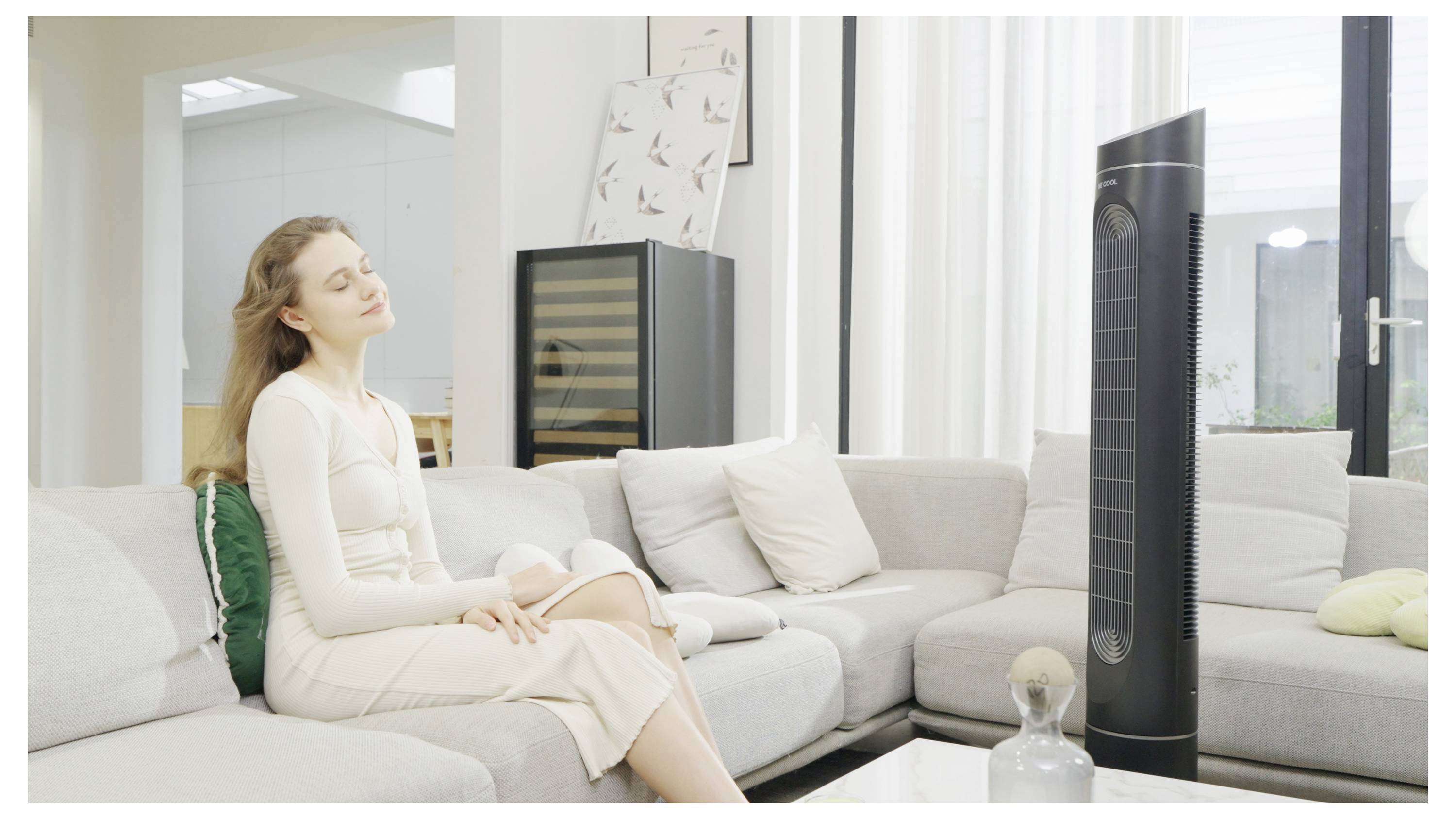 Eine Frau sitzt entspannt auf einem Sofa mit geschlossenen Augen und genießt die Brise eines großen, schwarzen Ventilators in einem modernen Wohnzimmer.