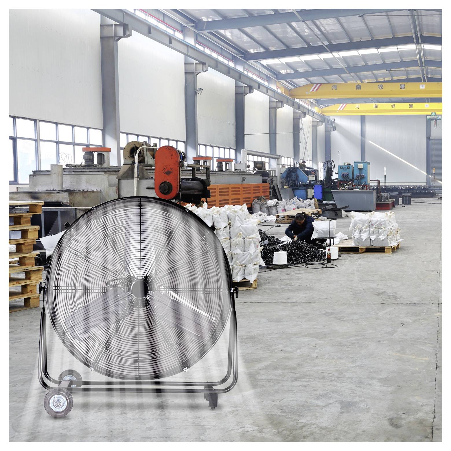 Ein großer Industrieventilator läuft in einem geräumigen Fabriklaghaus. Arbeiter und Maschinen befinden sich im Hintergrund, in der Nähe befinden sich Materialstapel.