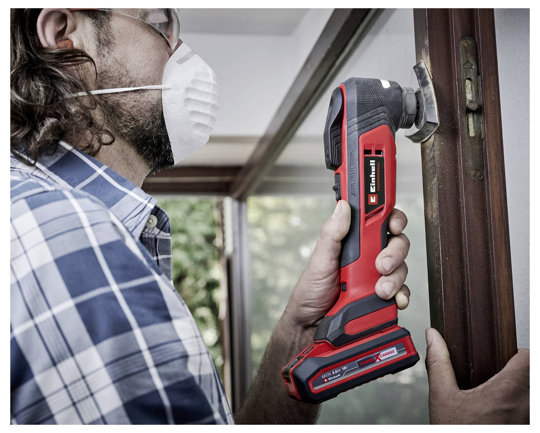 Eine Person mit einer Schutzbrille und einer Maske benutzt ein rotes Elektrowerkzeug, um an einem Holzrahmen neben einem Fenster zu arbeiten.