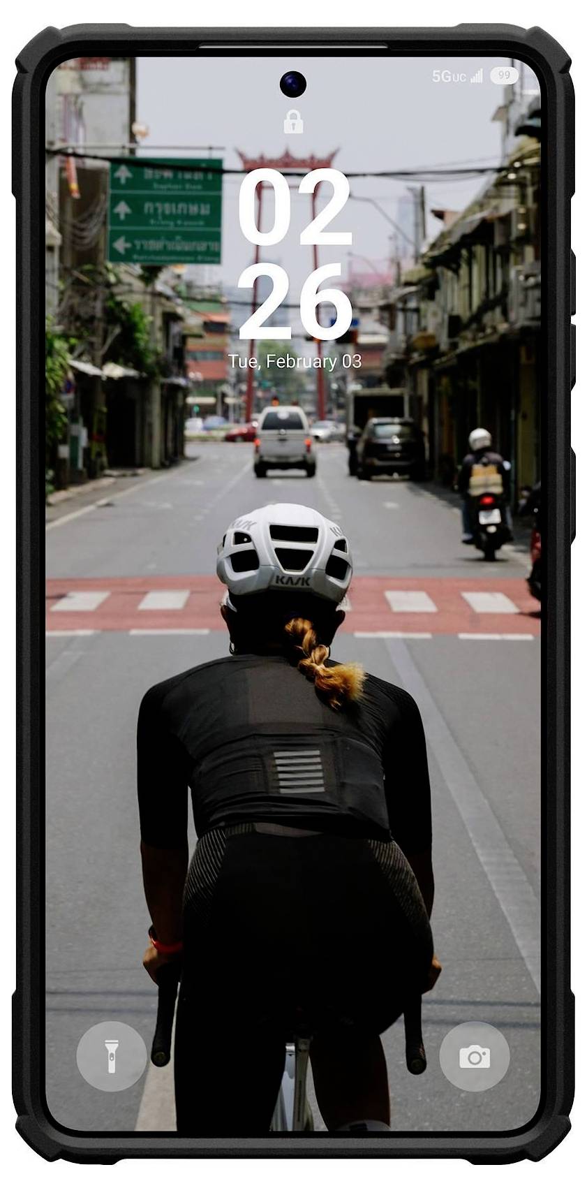 Eine Person auf einem Fahrrad fährt eine städtische Straße hinunter, von hinten betrachtet. Der Smartphonebildschirm zeigt „02:26 PM, Di., 3. Februar