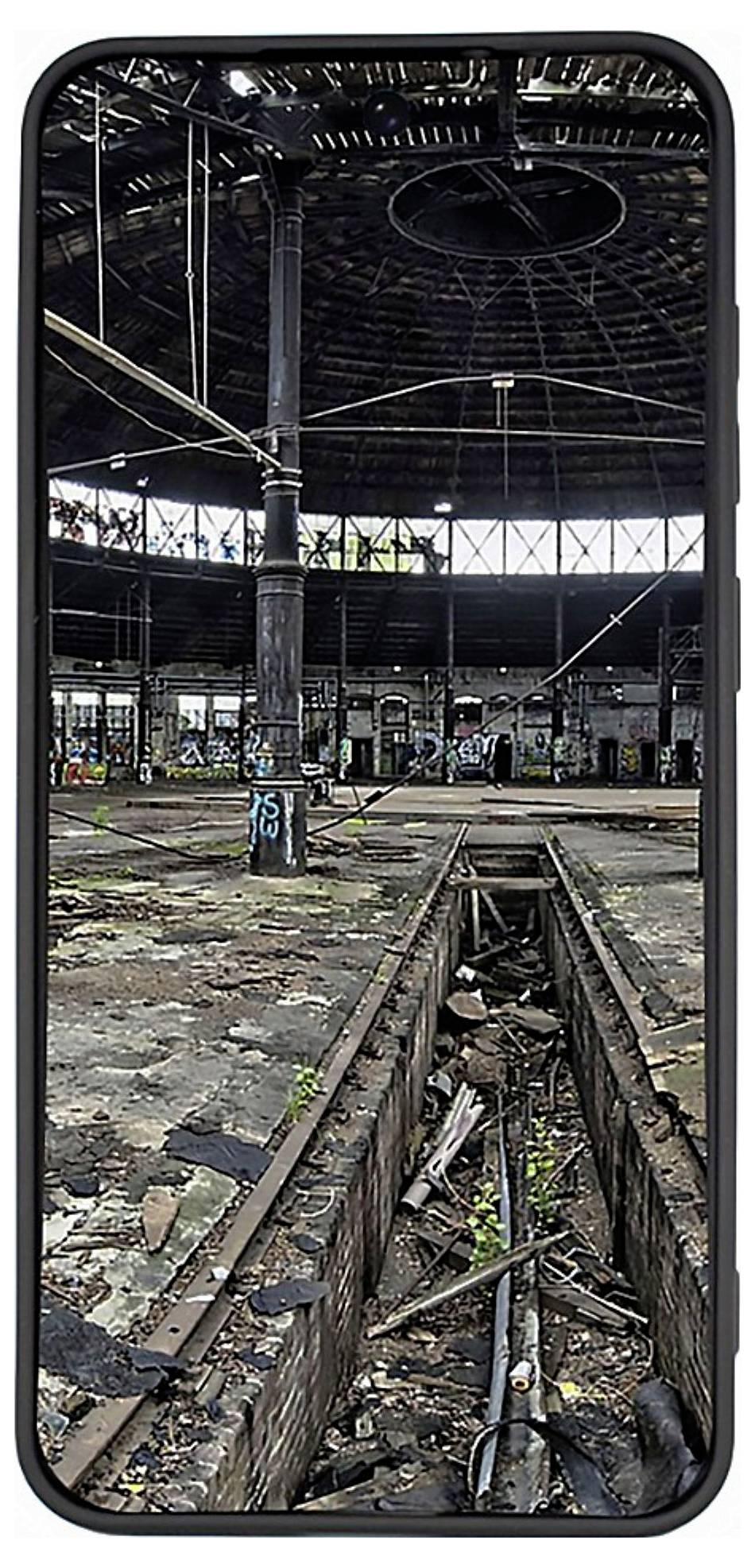 Ein verlassenes, mit Graffiti bedecktes Industriegebäude mit zerbrochenen Bodenfliesen und überwucherndem Unkraut, betrachtet durch einen Telefonbildschirmrahmen.