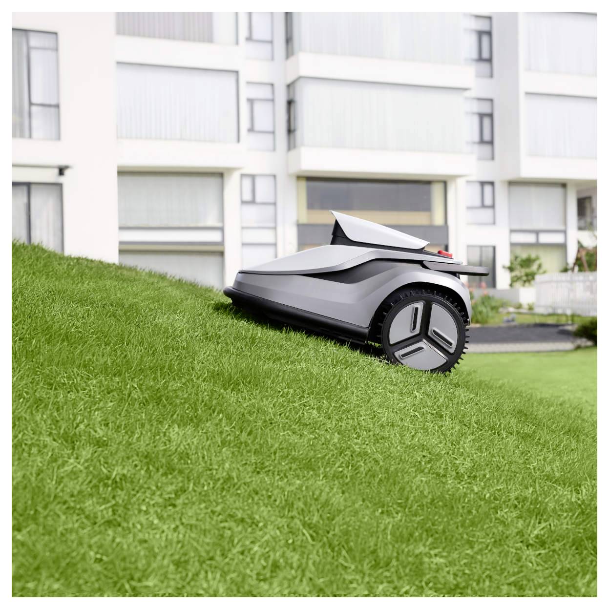 Ein Rasenroboter in Betrieb, der sanft einen Rasenhang hinuntergleitet, mit modernen Wohngebäuden im Hintergrund.