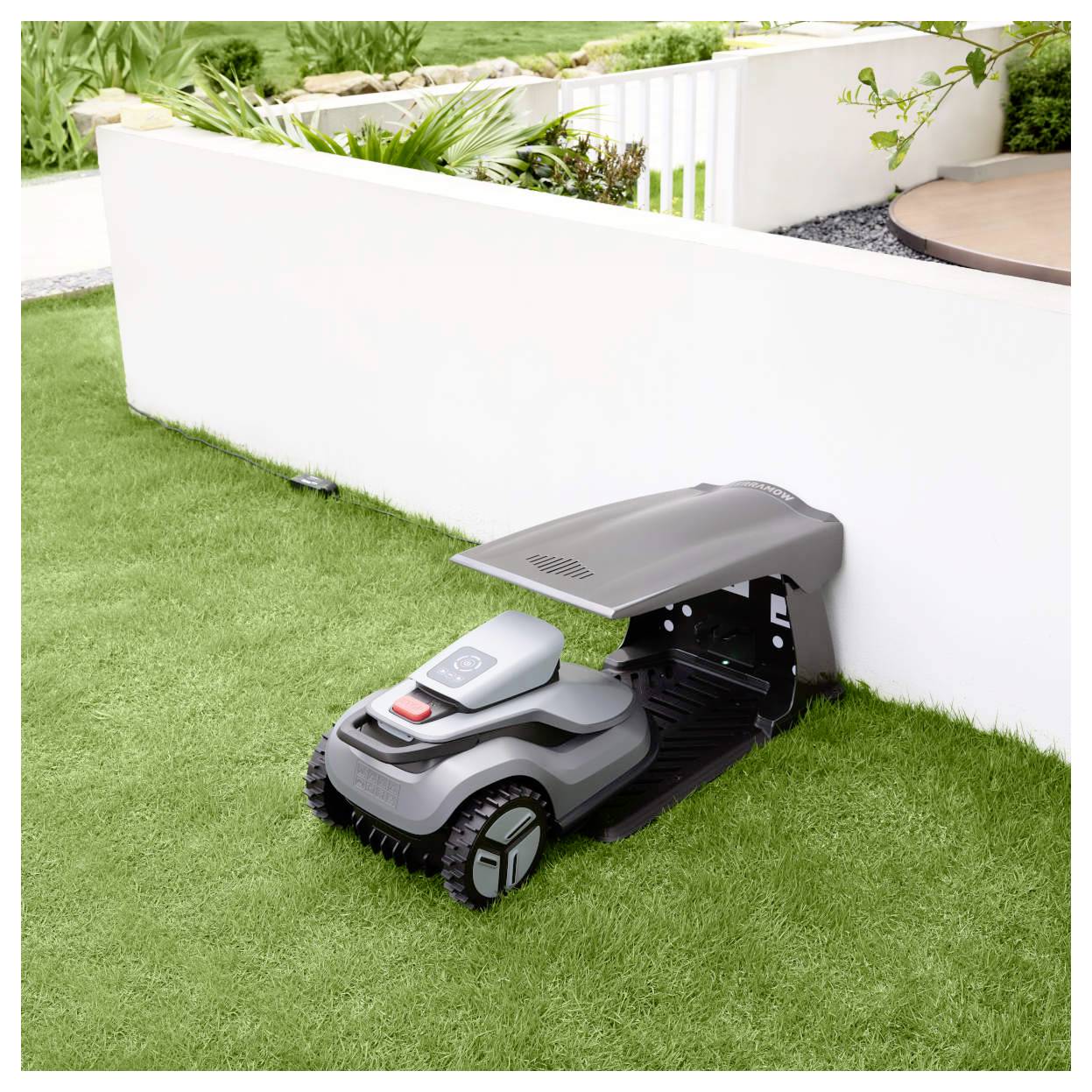 Ein Roboter-Rasenmäher ist unter einem kleinen Unterstand neben einer weißen Wand in einem Garten geparkt und zeigt sein kompaktes Design und seine Lagerlösung.
