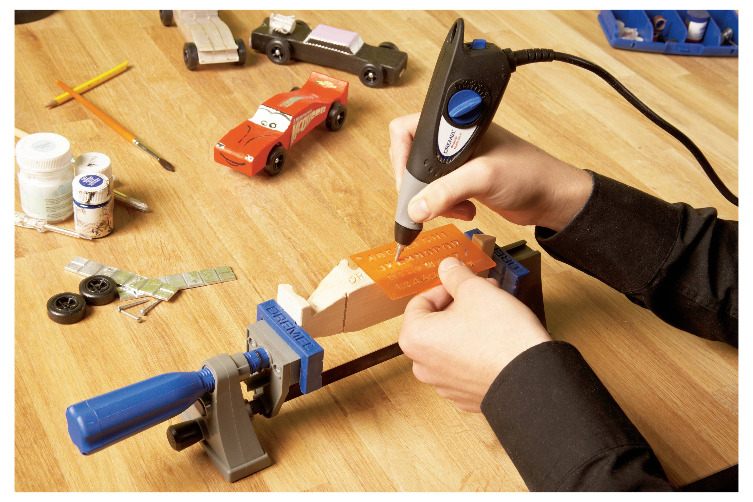 Eine Person nutzt ein Rotationswerkzeug, um ein Modell-Holzauto zu schnitzen, das in einem Schraubstock auf einem Werkstatttisch befestigt ist, umgeben von Farben und Modellteilen.