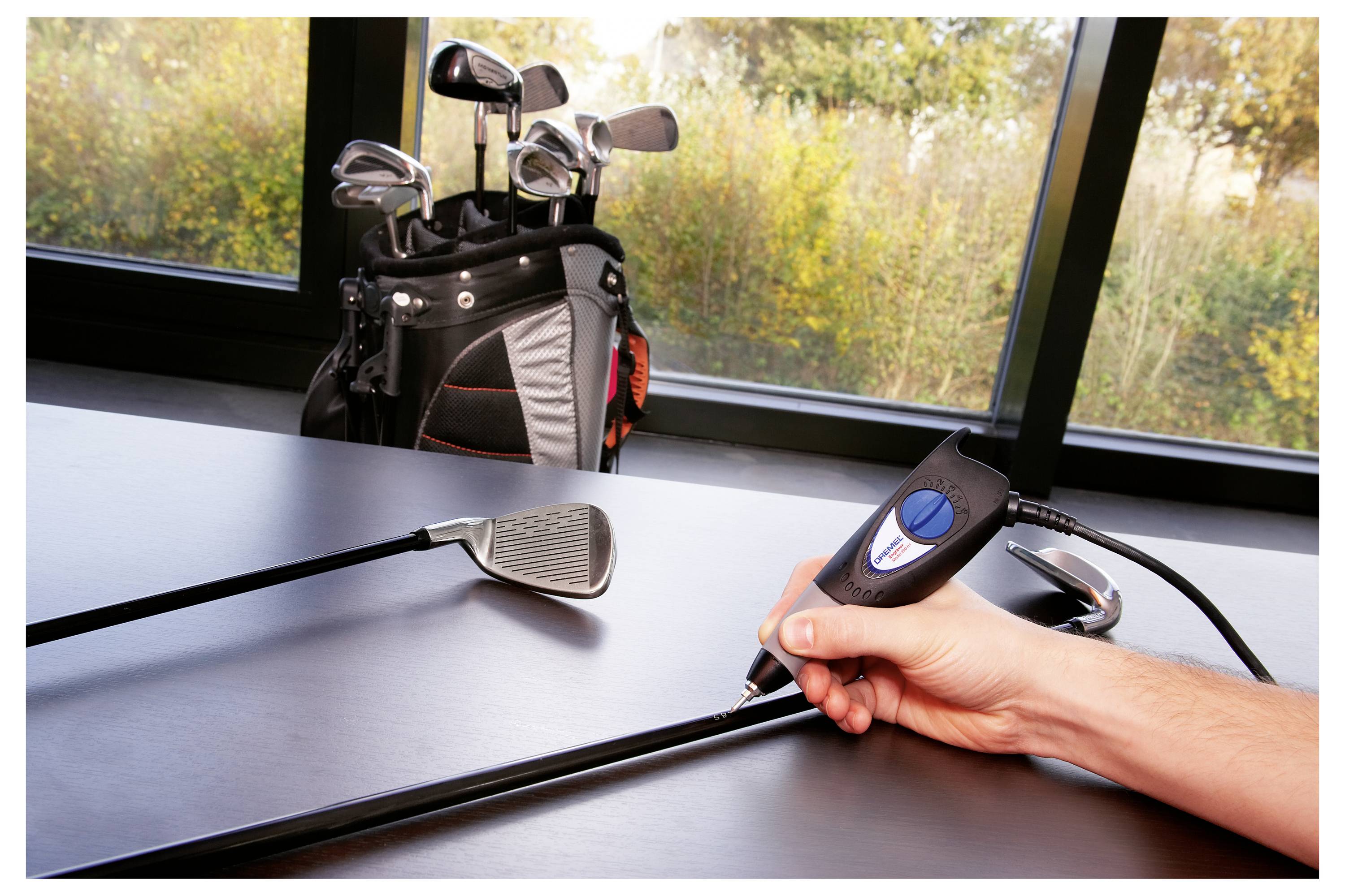 Eine Person graviert einen Golfschläger-Schaft mit einem tragbaren Werkzeug, während eine Golftasche mit Schlägern im Hintergrund in der Nähe eines großen Fensters steht.