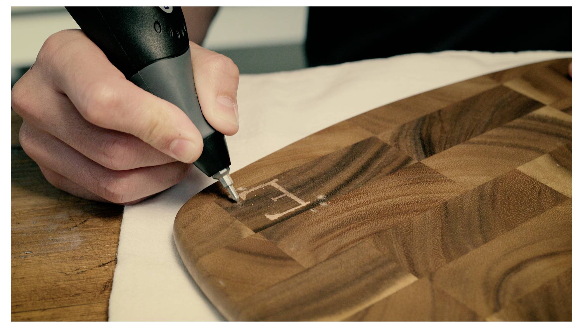 Hand mit einem Werkzeug, das den Buchstaben 'E' auf eine Holzoberfläche ätzet, wobei ein Handwerk oder Holzbearbeitungsprojekt in Arbeit demonstriert wird.