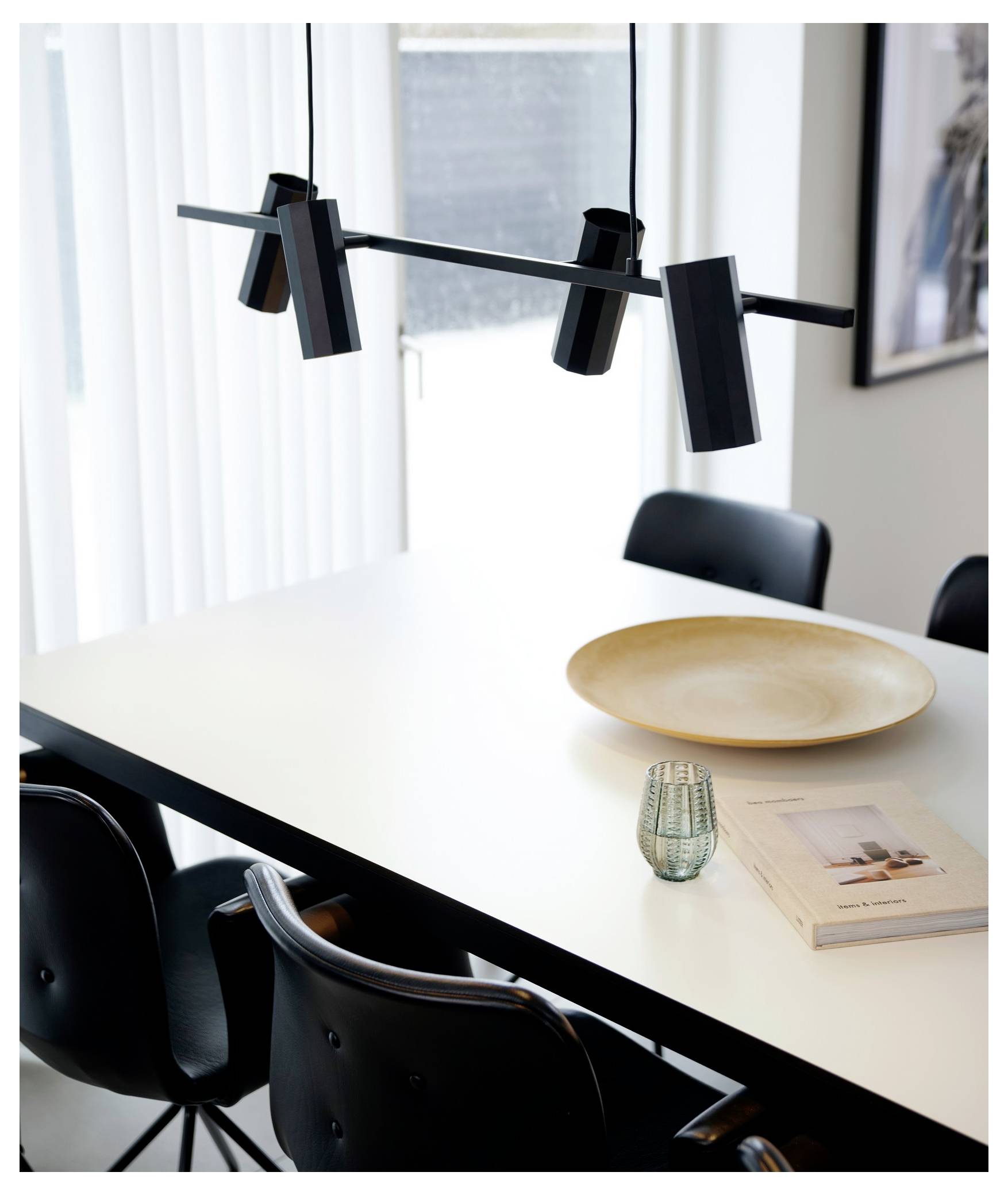 Ein moderner Speisebereich mit einem weißen Tisch, schwarzen Stühlen, schwarzer Hängeleuchtenkonstruktion, Keramikschüssel, kleiner Vase und zwei Büchern.