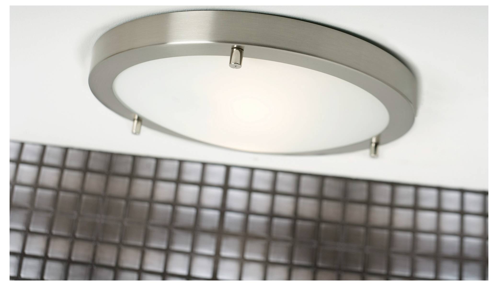 Eine moderne Deckenleuchte mit einer runden, gebürsteten Metallfassung und Mattglas, montiert über einem Raster aus kleinen quadratischen Fliesen.