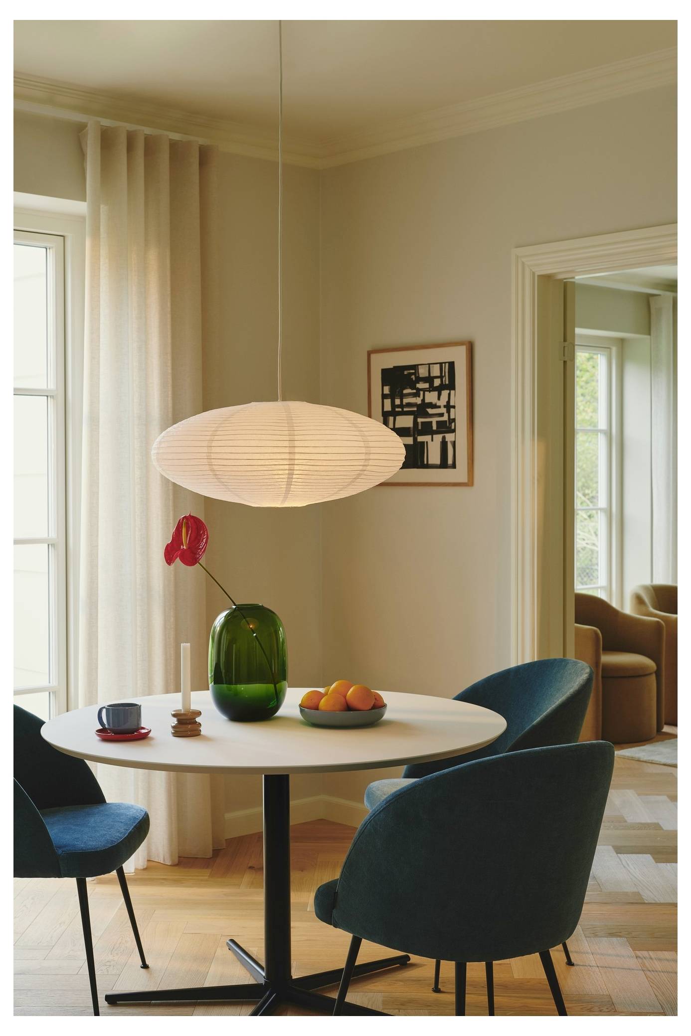 Ein runder Esstisch mit dunkelblauem Stühlen, eine grüne Vase mit einer roten Blume, eine Schale mit Orangen und eine hängende weiße Lampe in einem gemütlichen Zimmer.