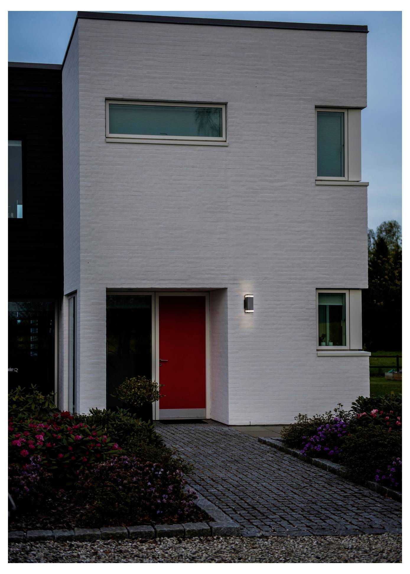 Modernes zweistöckiges Haus mit weißer Backsteinfront, roter Haustür und rechteckigen Fenstern. Ein Backsteinweg führt zum Eingang.