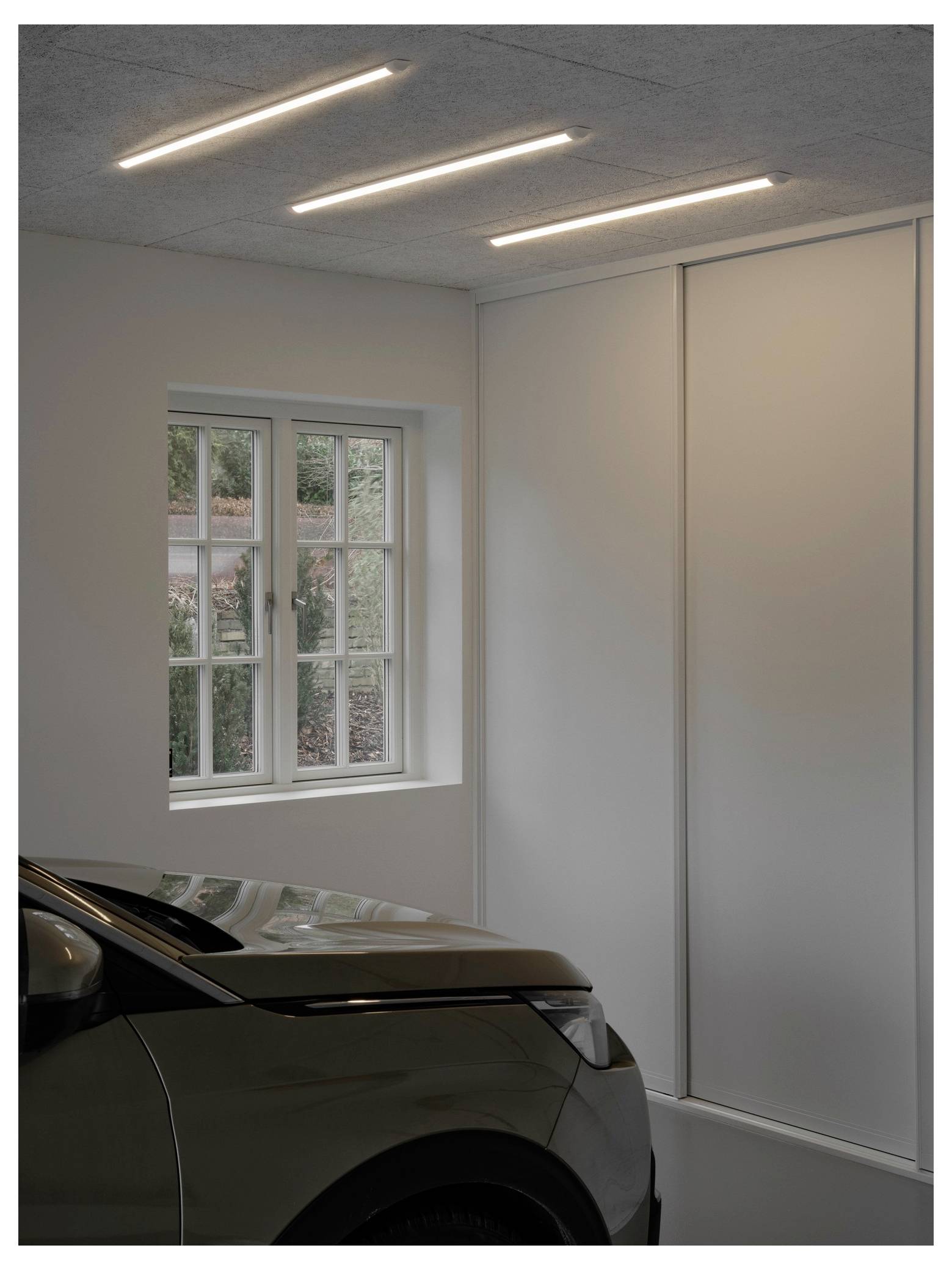 Ein Auto ist in einer modernen Garage mit einem Fenster auf der linken Seite und drei linearen Deckenleuchten geparkt.