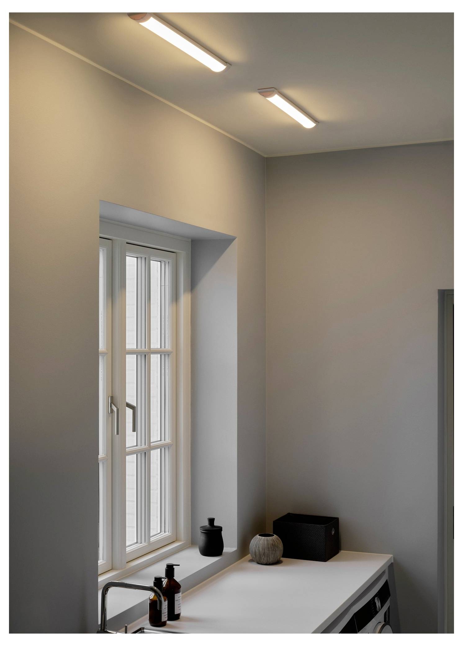 Eine minimalistische Küchennische mit einem Waschbecken unter einem Fenster. Zwei lineare Deckenleuchten beleuchten den Raum. Dekorative Gegenstände stehen auf der Arbeitsplatte.
