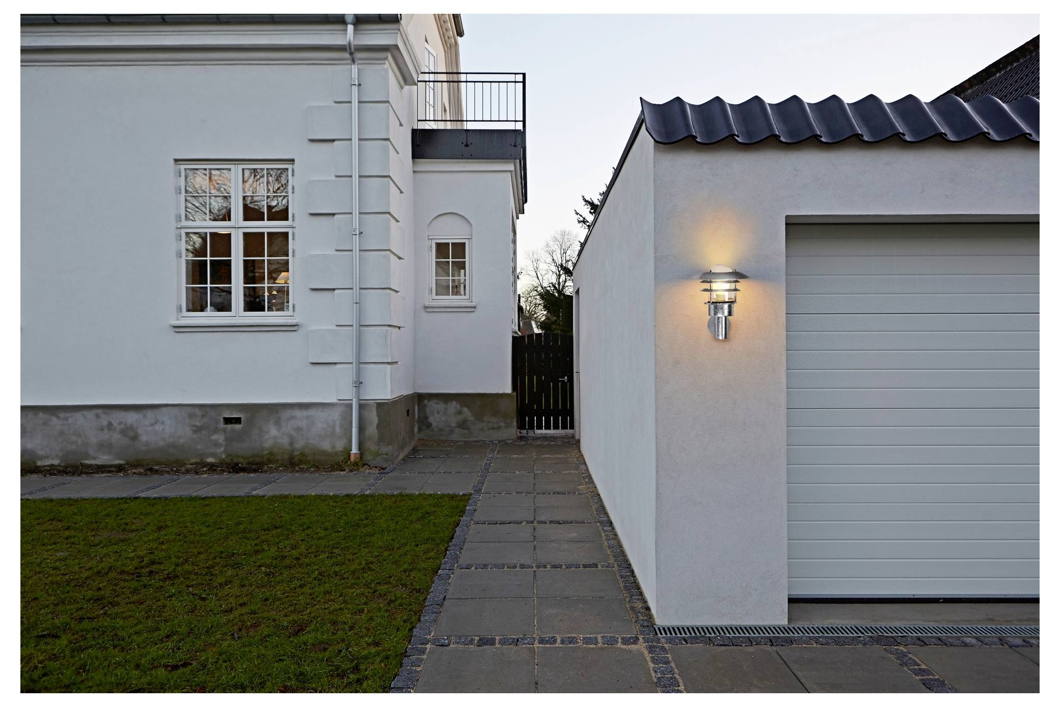 Ein weißes Haus mit einer Garage auf der rechten Seite, beleuchtet durch ein Außenlicht. Der Weg ist gepflastert, und auf der linken Seite befindet sich eine kleine Rasenfläche.