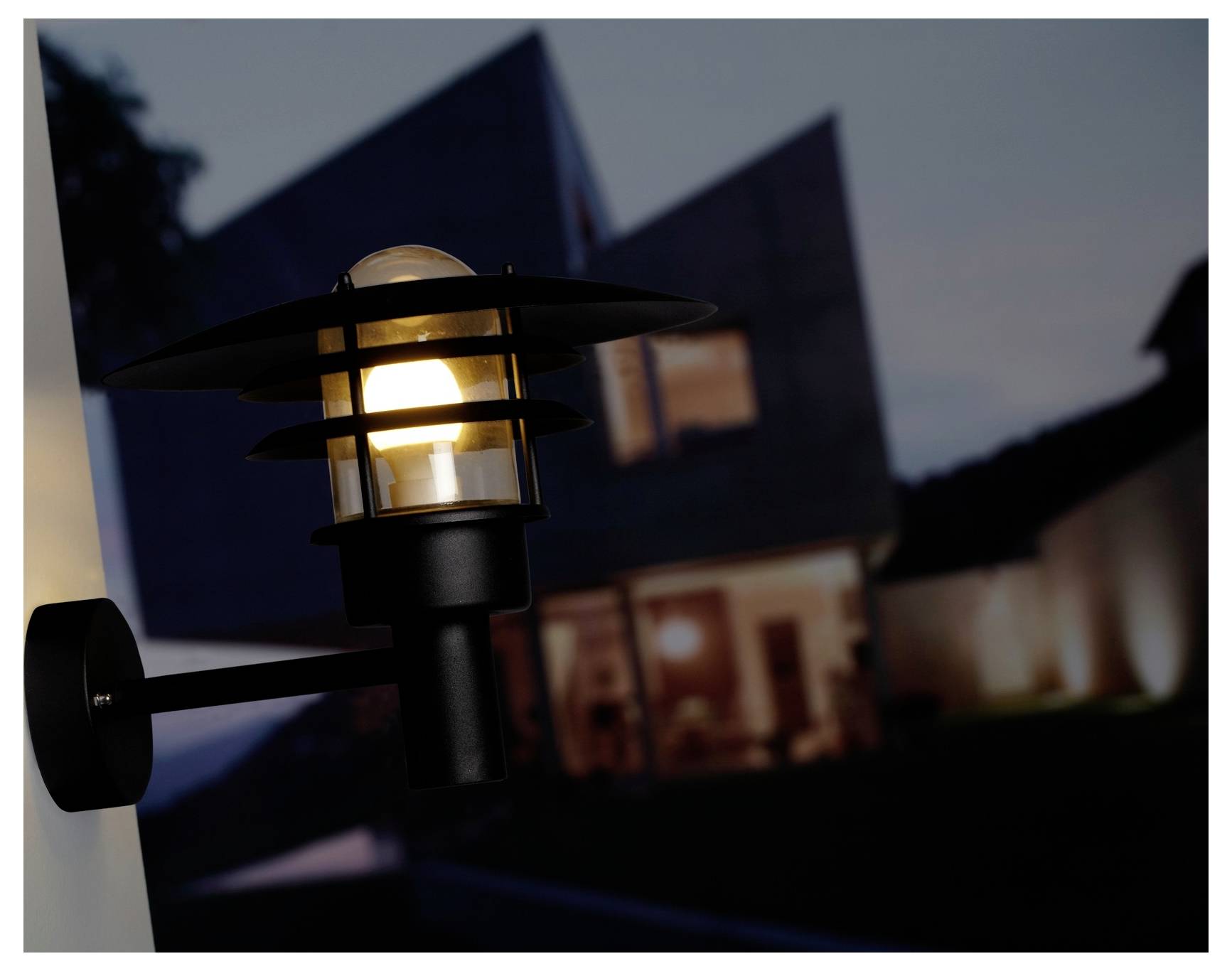 Eine moderne Außenwandleuchte ist gegen die dunkle Kulisse eines zeitgenössischen Hauses in der Dämmerung erleuchtet.