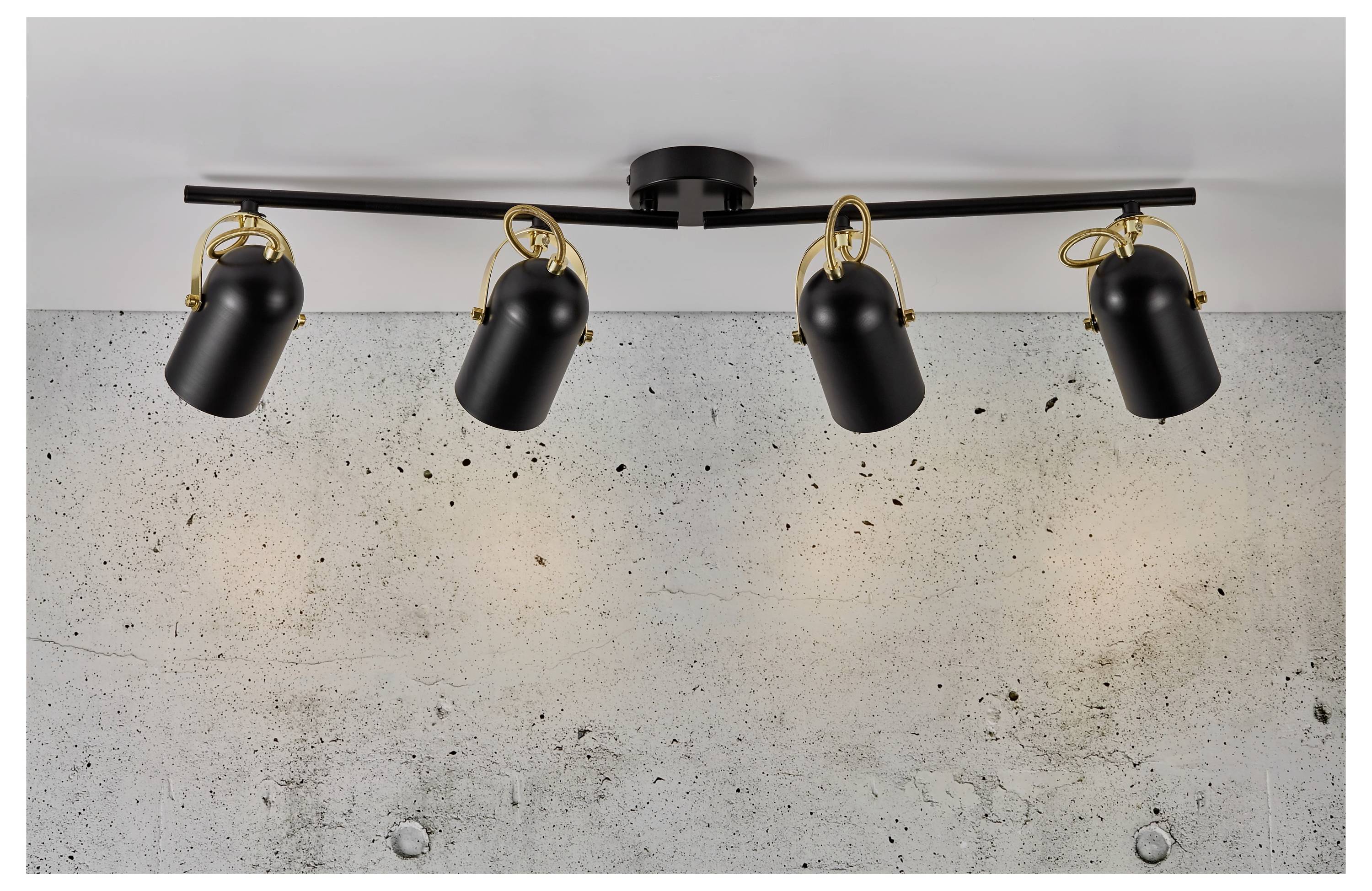 „Deckenleuchte mit vier verstellbaren schwarzen Spotlights, montiert auf einer schwarzen Stange mit dekorativen Goldringen, vor einer Betonwand.