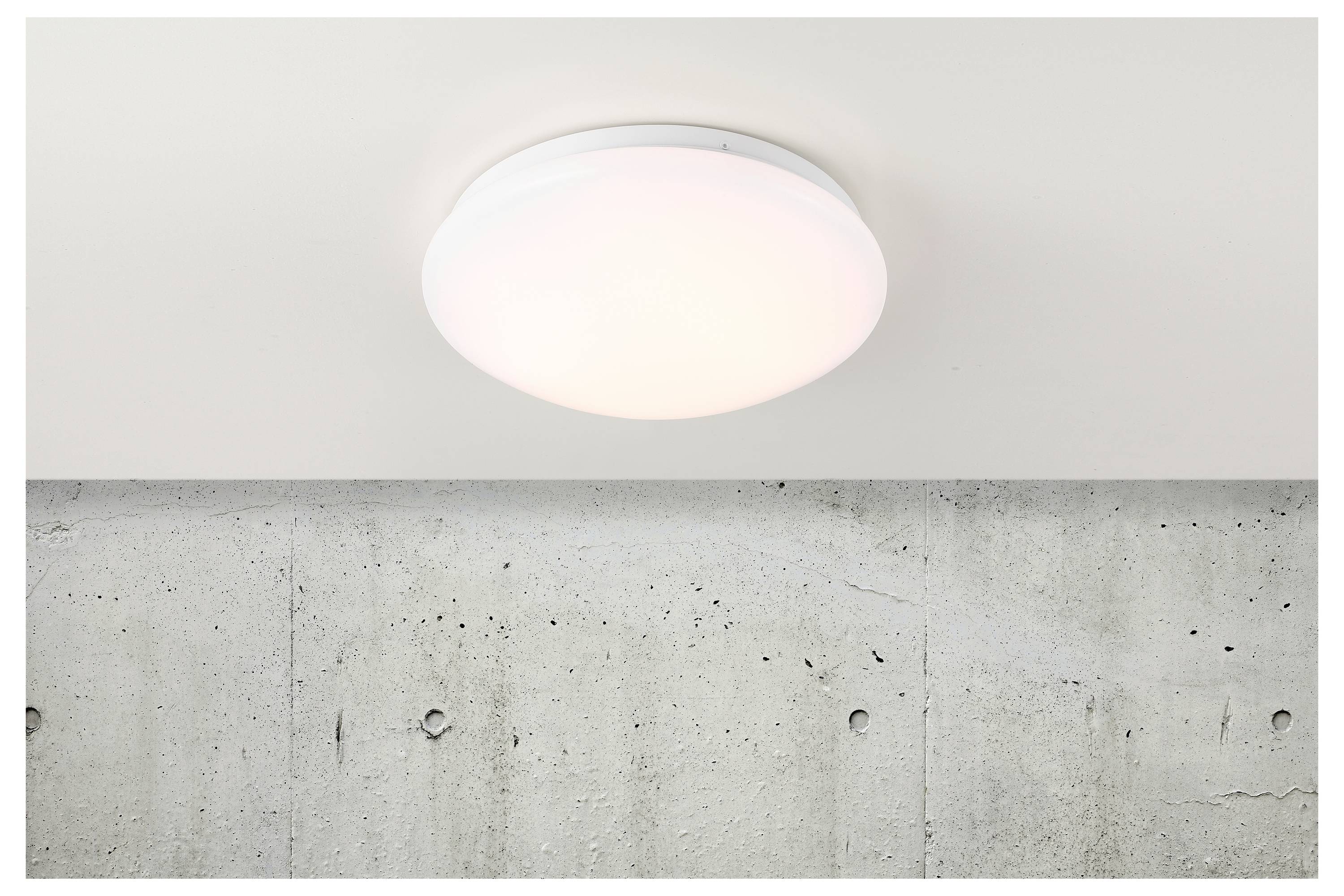 Eine runde, weiße Deckenleuchte ist auf einer glatten, hellfarbigen Decke über einer Betonwand montiert.